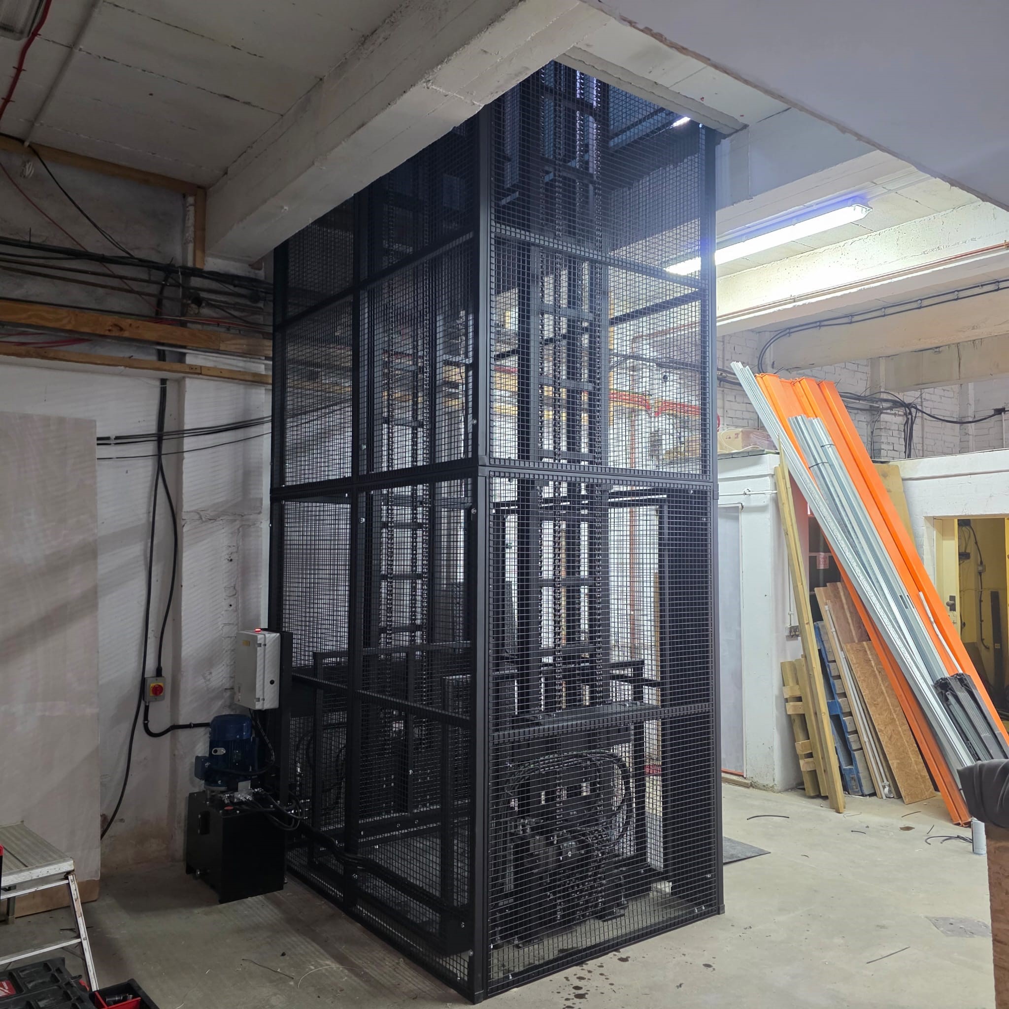MezzHD 1000kg Mezzanine Goods Lift. A goods lift with a black metal mesh cage is installed in an unfinished industrial space, with exposed wiring, pipes, and building materials visible around it.