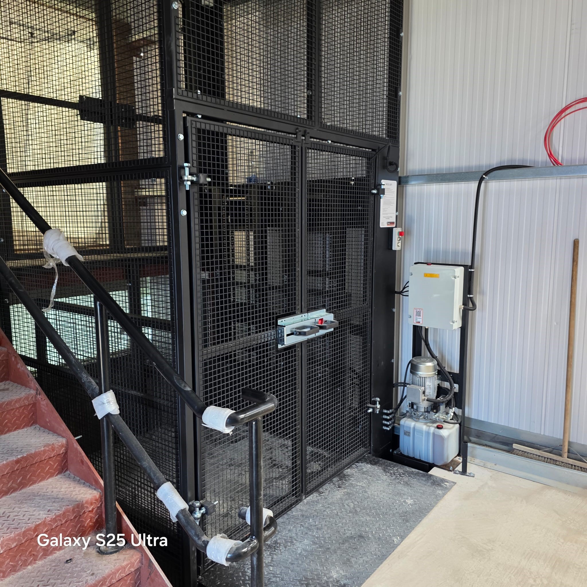 A black metal industrial lift with mesh doors is installed next to a red staircase and a white wall. Electrical wiring and a control box are mounted nearby. The words Galaxy S25 Ultra appear in the bottom left corner.