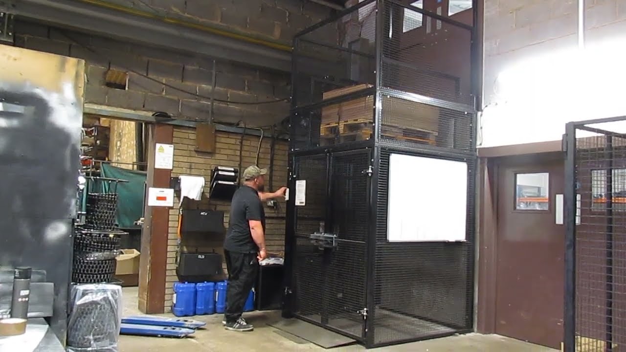 A person in work clothes operates a vertical platform lift inside an industrial warehouse, surrounded by metal mesh cages, shelves, and various equipment.
