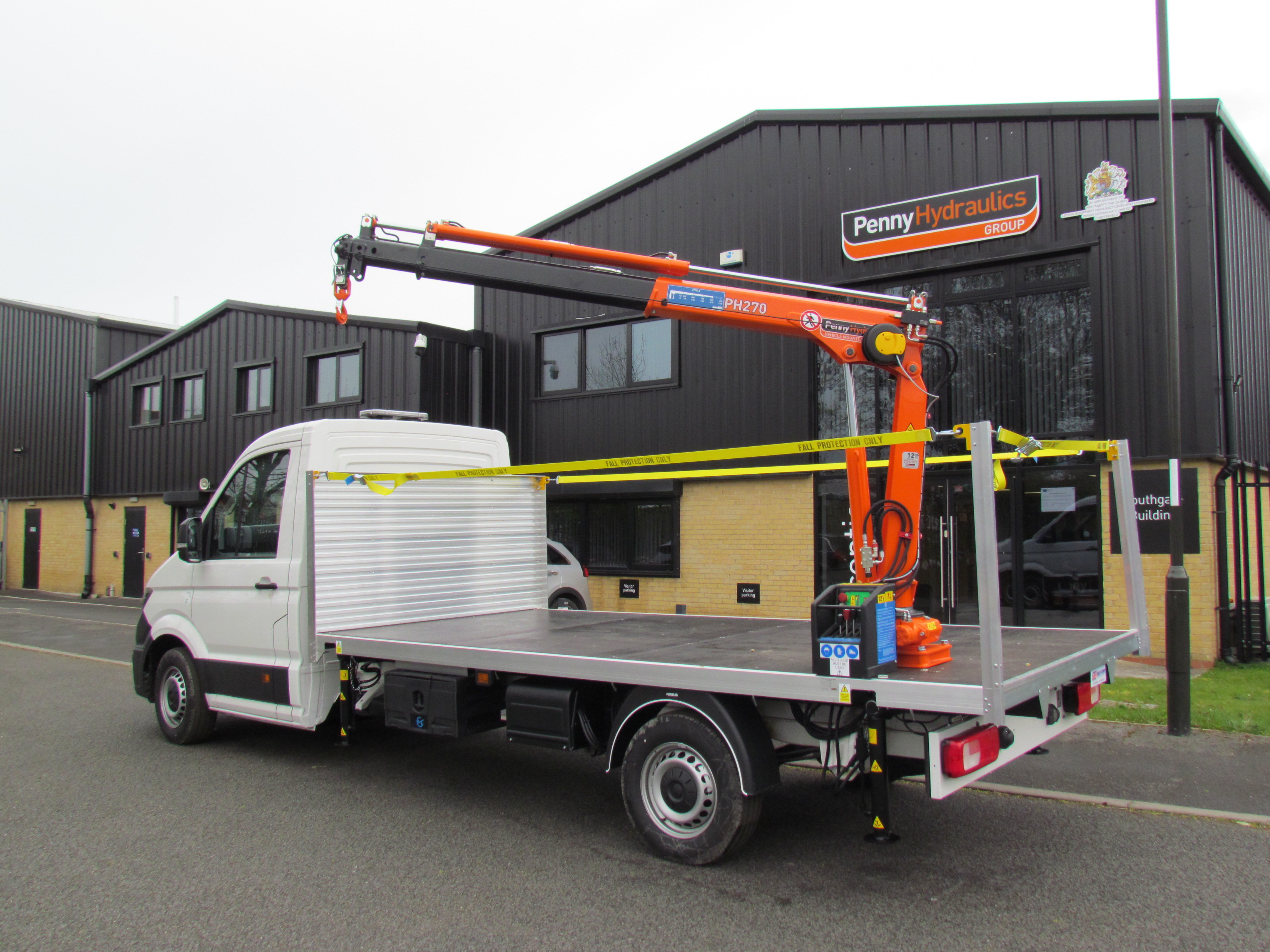 A white flatbed lorry with an orange hydraulic crane mounted on its bed is parked outside a black industrial building labelled Penny Hydraulics. The crane arm is extended over the lorry.