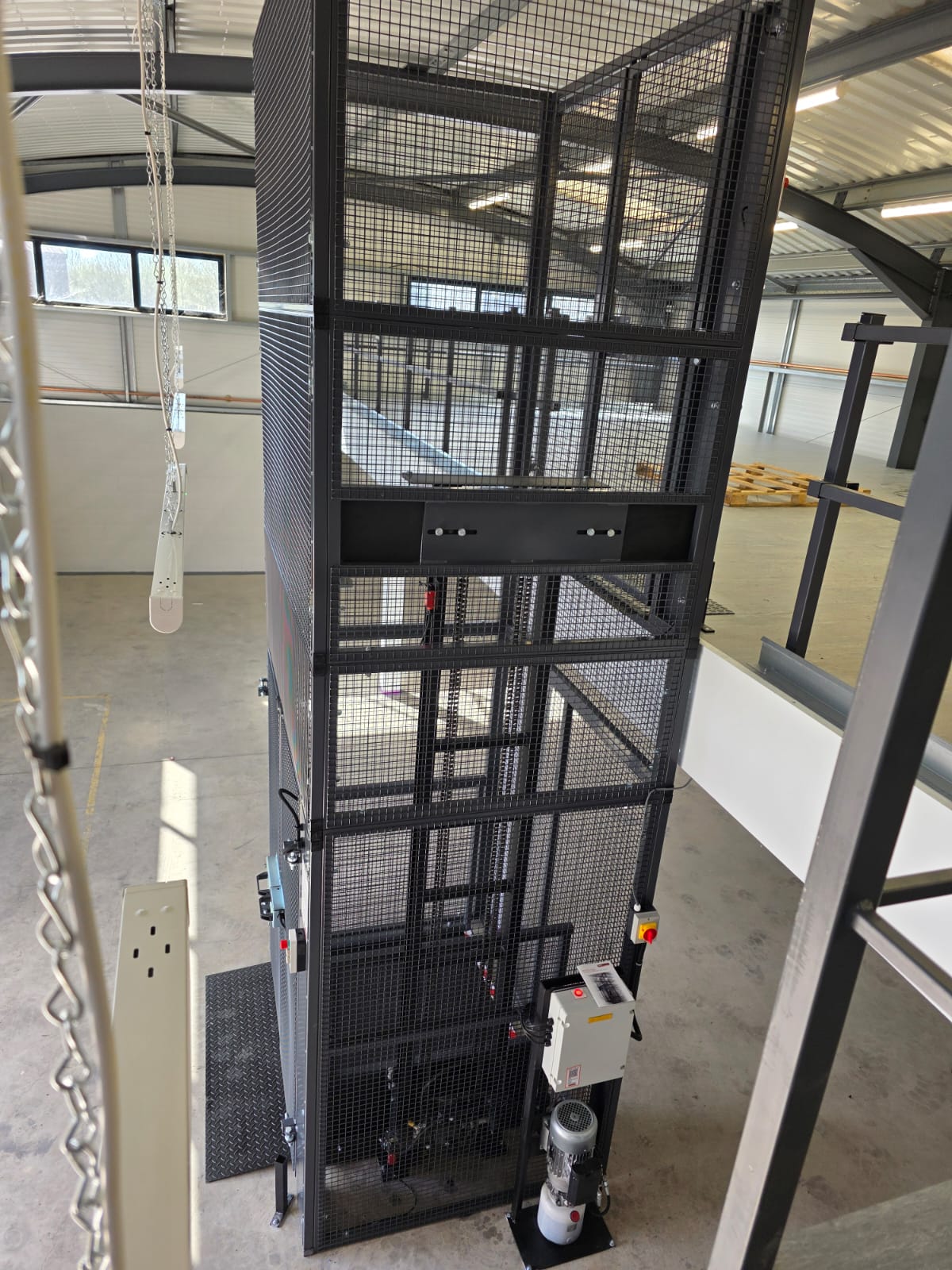 An empty industrial lift shaft with metal railings and beams, viewed from above. The structure is enclosed by glass panels and the background shows a building interior.