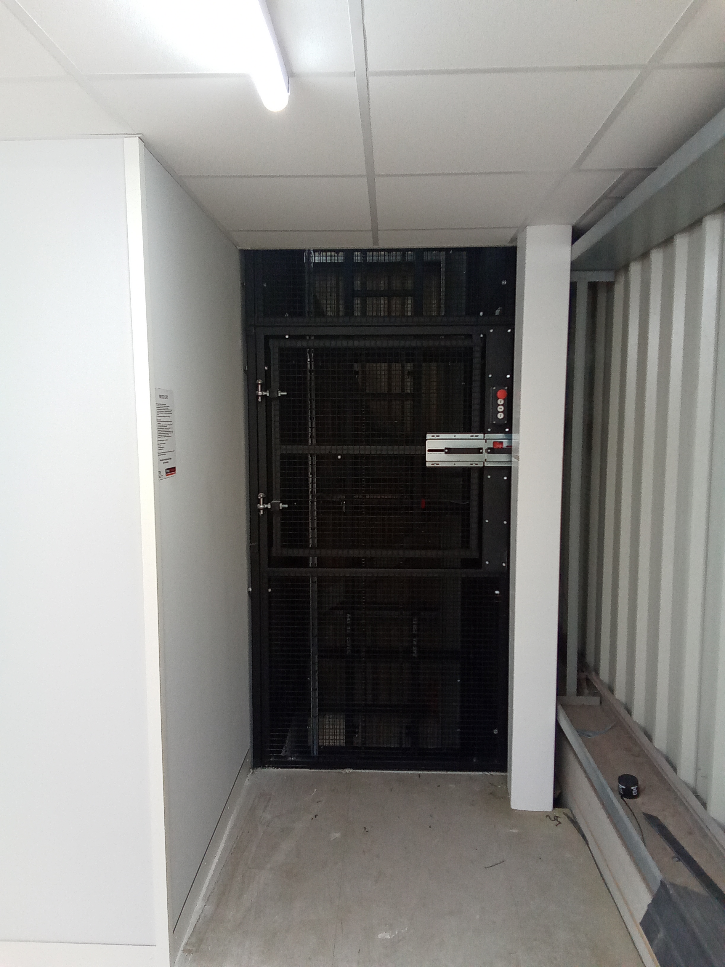 A small goods or service lift with a black metal gate is located at the end of a narrow corridor with white walls and a tiled ceiling. The floor is concrete and fluorescent lighting is overhead.