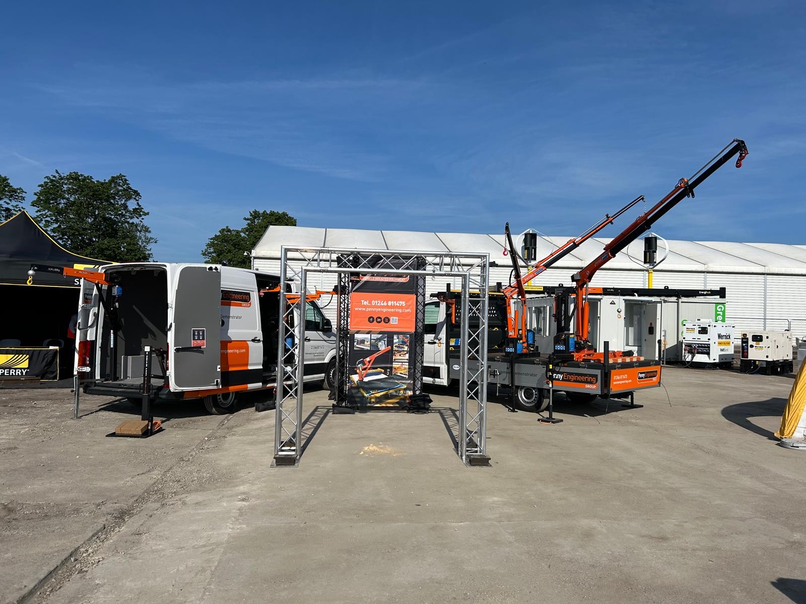 Outdoor exhibition display featuring utility vehicles, a van, and lorries with mounted cranes. A metal frame holds a banner at the centre, with company branding and information visible. Blue sky and white marquees are in the background.