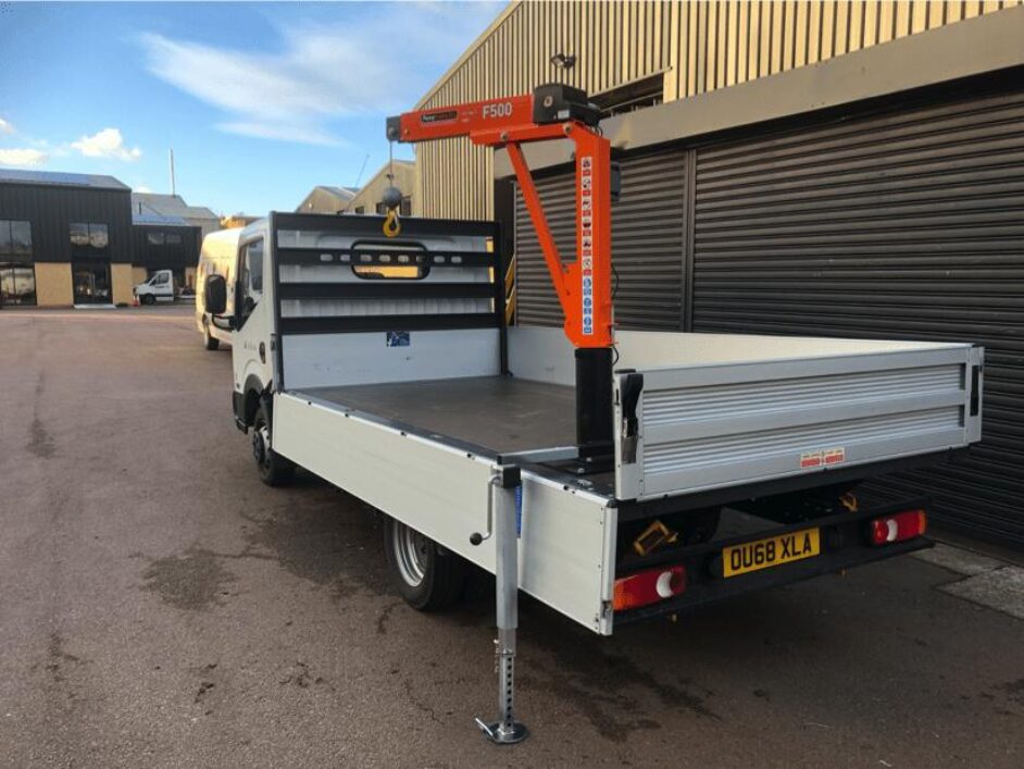 A white flatbed lorry with an orange F500 crane mounted on the back is parked outside industrial buildings on a paved area. The lorry bed is empty, and the crane's support leg is extended to the ground.