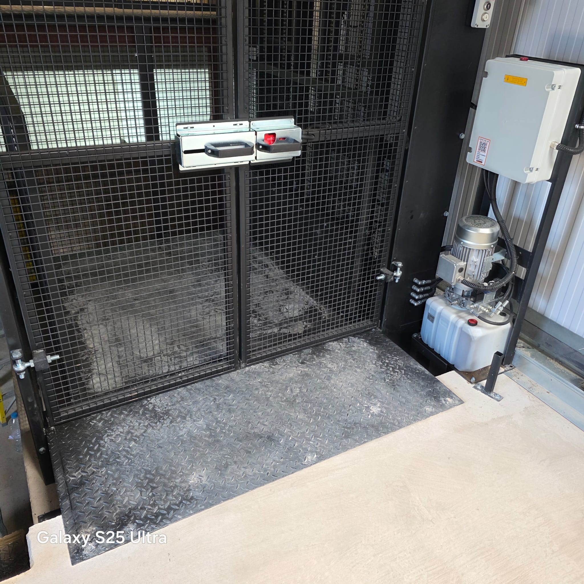 A metal gate with a locking mechanism is installed on a grated platform with a chequer plate floor. A hydraulic power unit is mounted nearby. The area appears to be part of an industrial or warehouse facility.