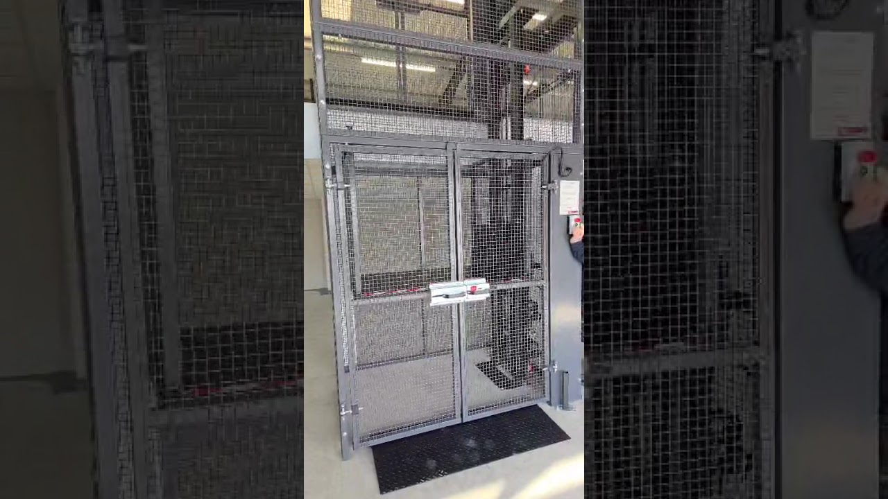 A person presses a button to operate a large metal safety cage with mesh walls and a gate inside an industrial or warehouse setting. The cage appears to be used for secure entry or equipment access.