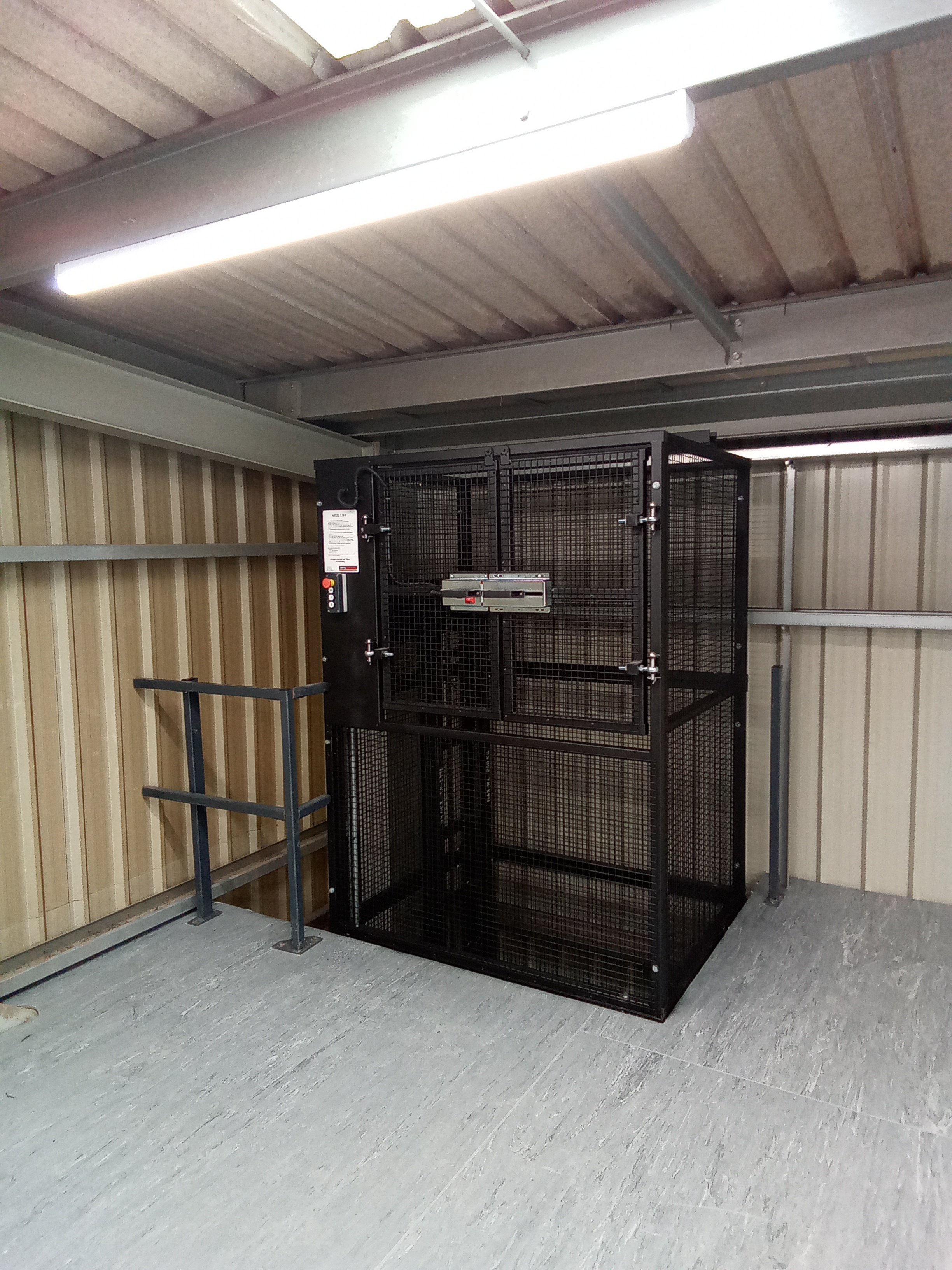 A black metal cage lift is installed on a mezzanine floor with metal railings and corrugated wall panels; fluorescent lighting is overhead.