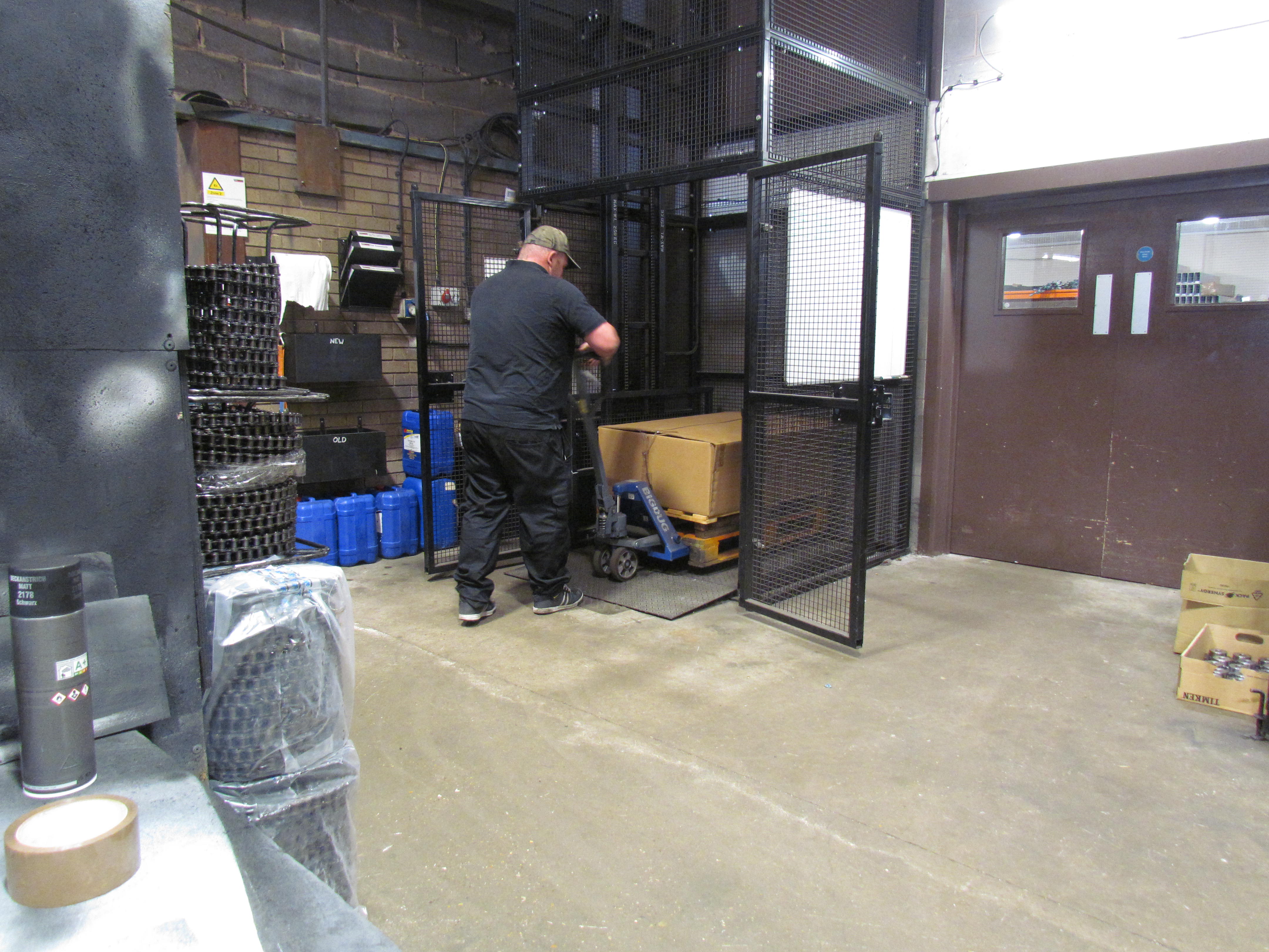 A person moves a large cardboard box on a pallet truck inside a gated storage area in an industrial warehouse, surrounded by shelves with blue containers and various supplies.