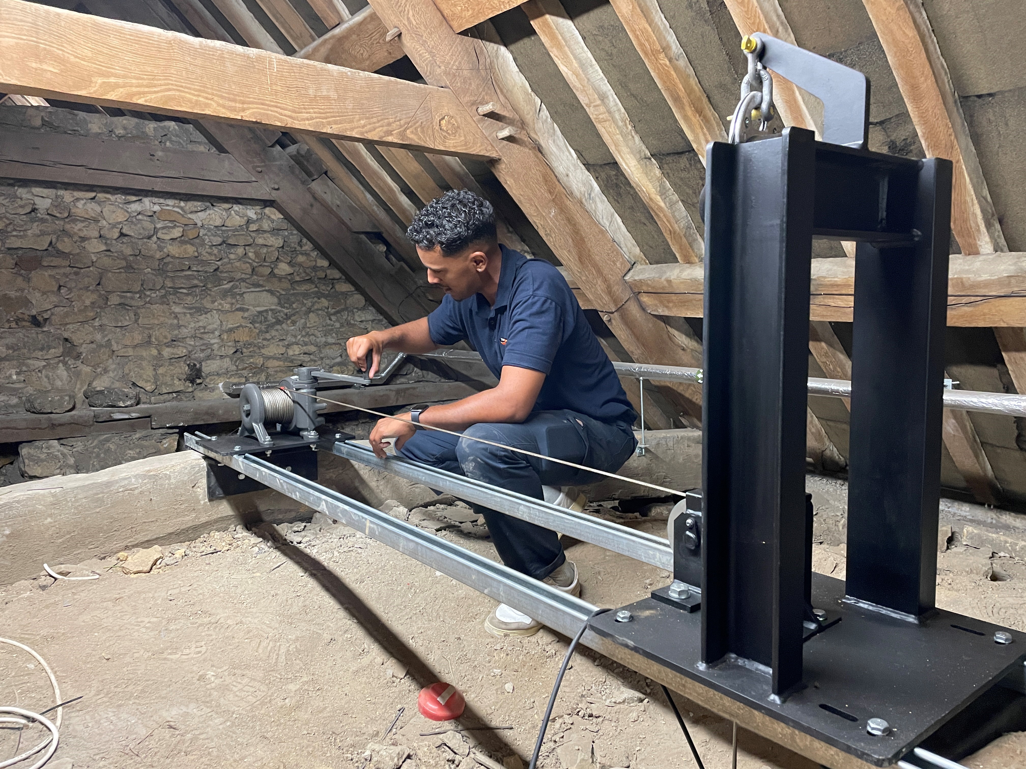 A man crouches in a loft, working with a metal winch system mounted on rails among wooden beams and stone walls. He appears to be installing or adjusting cables on the equipment.