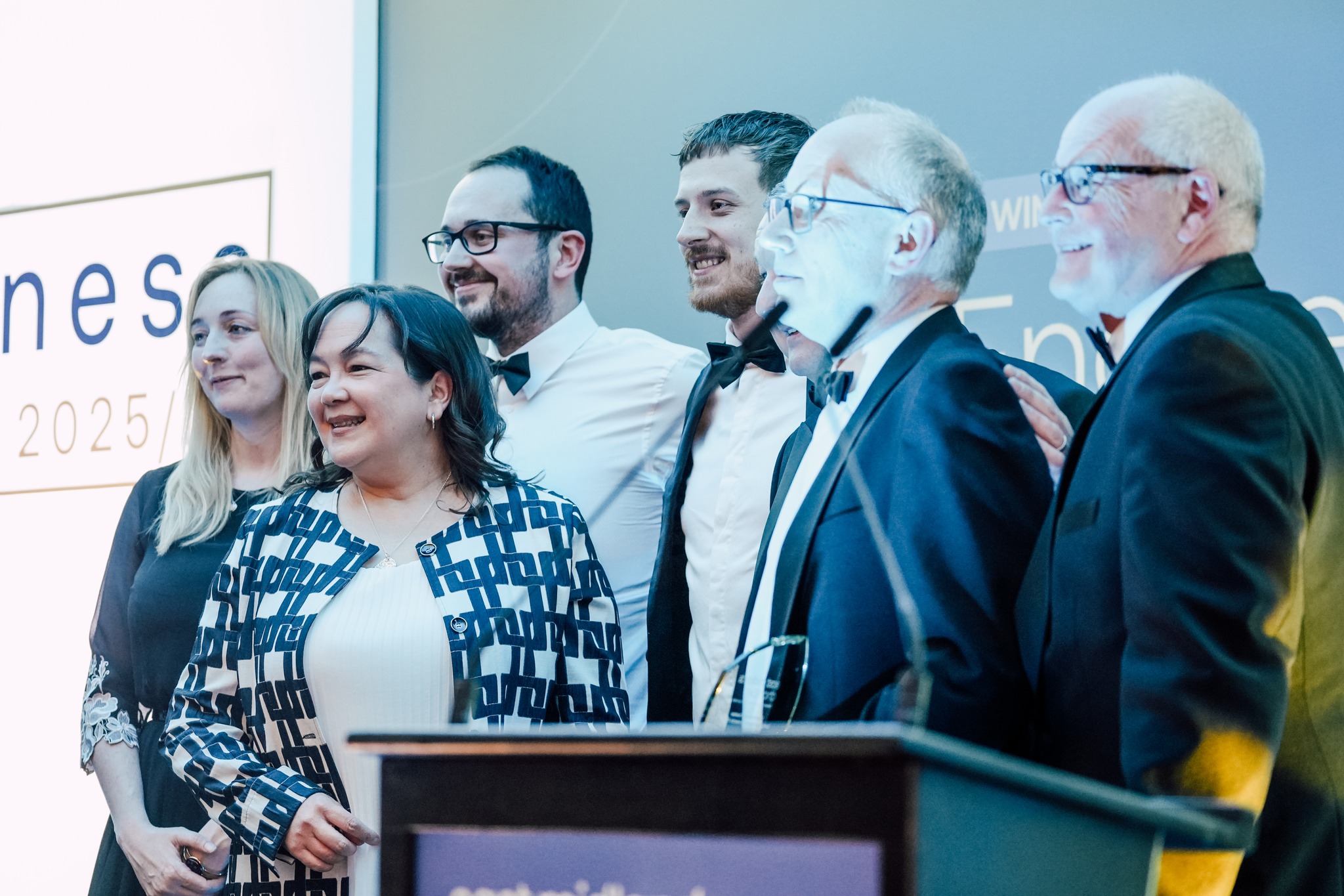 Six people dressed formally are smiling and posing together for a group photo at an indoor event, with a lectern and a partially visible sign in the background.