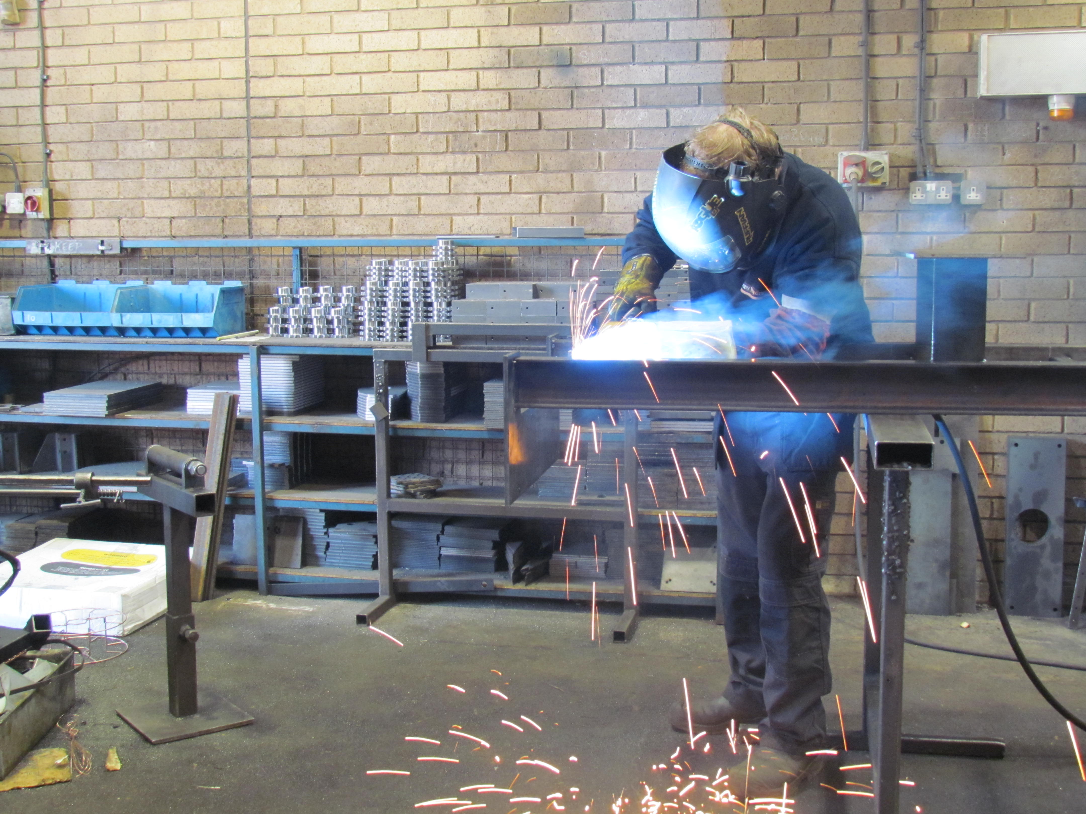 A person wearing protective gear and a welding helmet is welding metal in a workshop, with bright sparks flying around. Shelves with metal parts and equipment are visible in the background.
