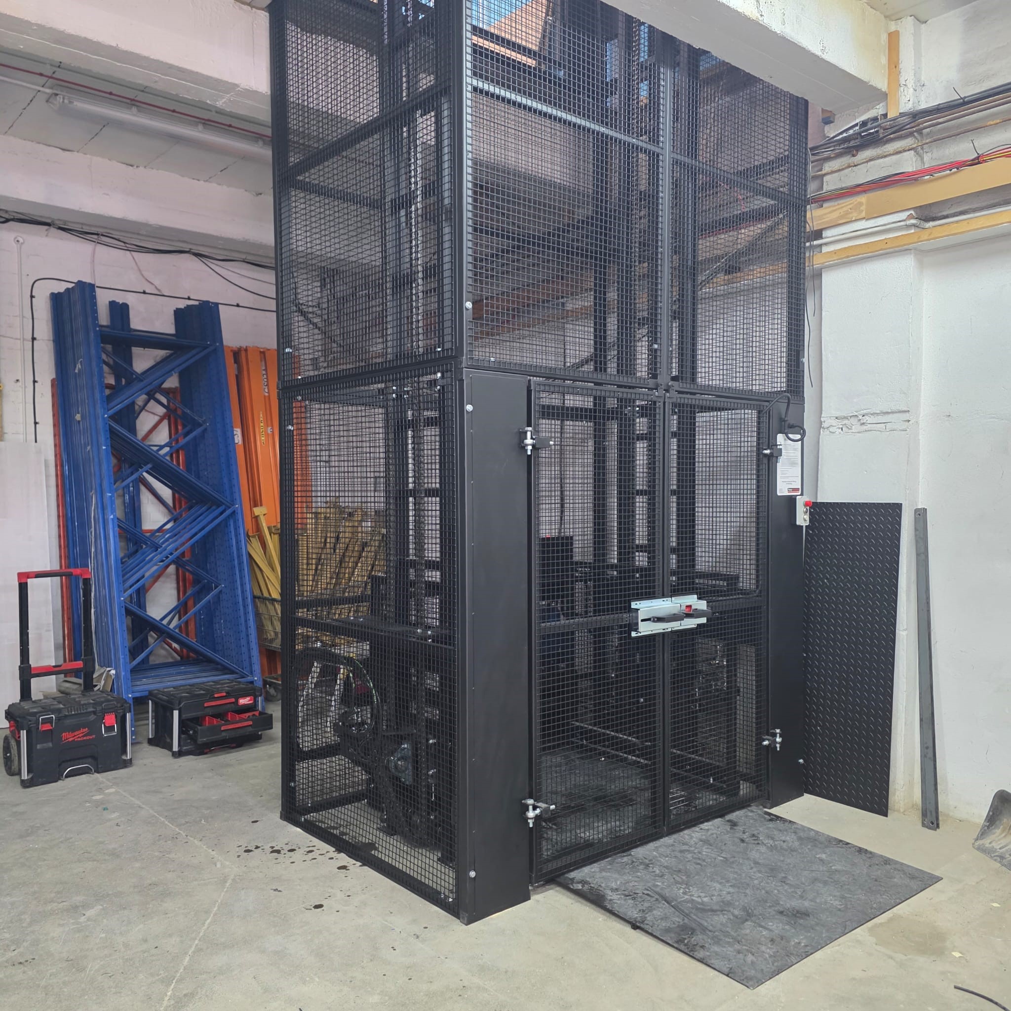 MezzHD 1000kg Mezzanine Goods Lift. A black metal industrial lift cage with mesh sides stands in a workshop, surrounded by tools, equipment, and building materials. Blue and orange racks and a red tool case are nearby.