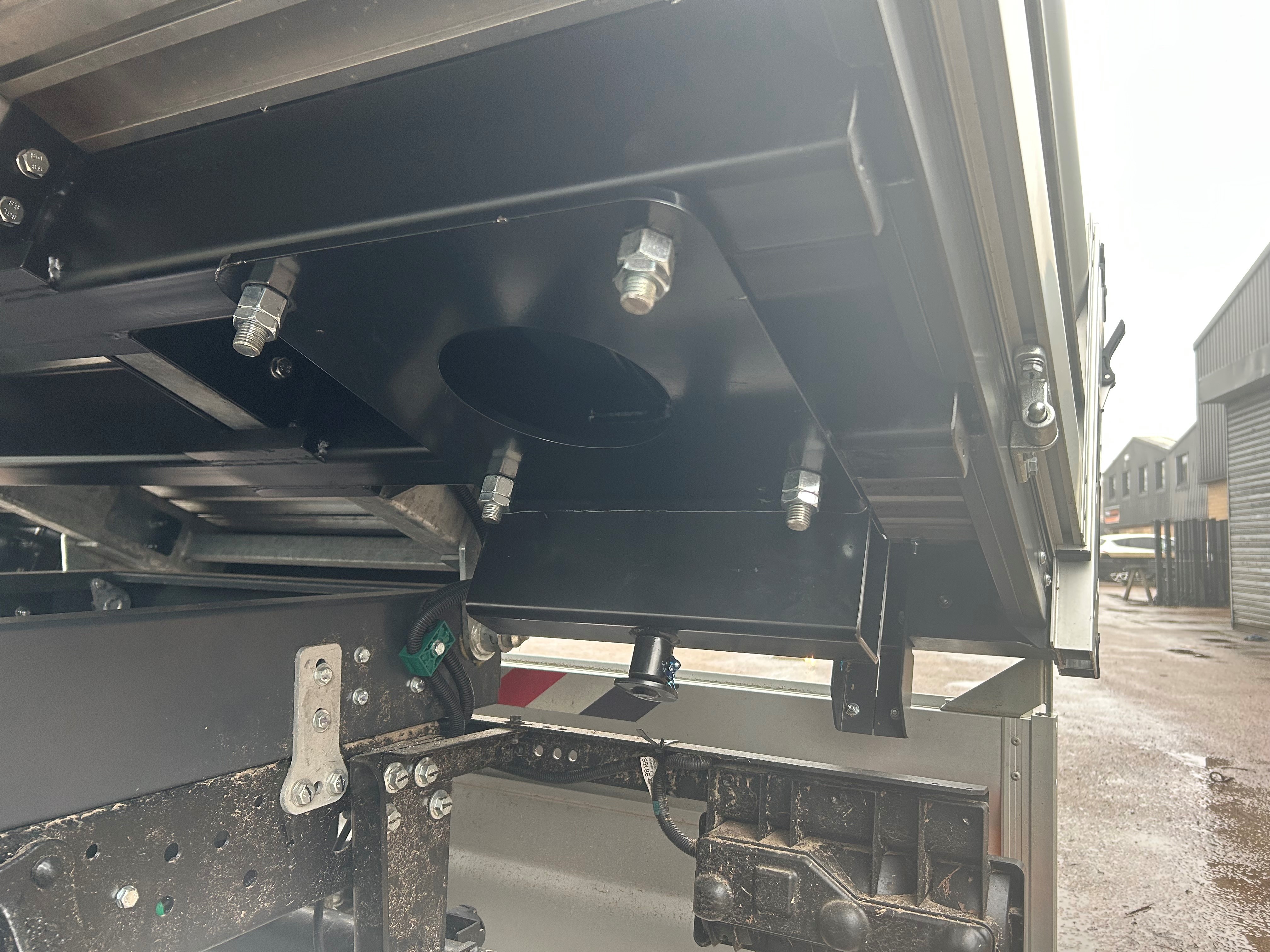 Close-up view of the underside of a lorry bed showing metal brackets, bolts, and mechanical components, with a building and wet ground visible in the background.
