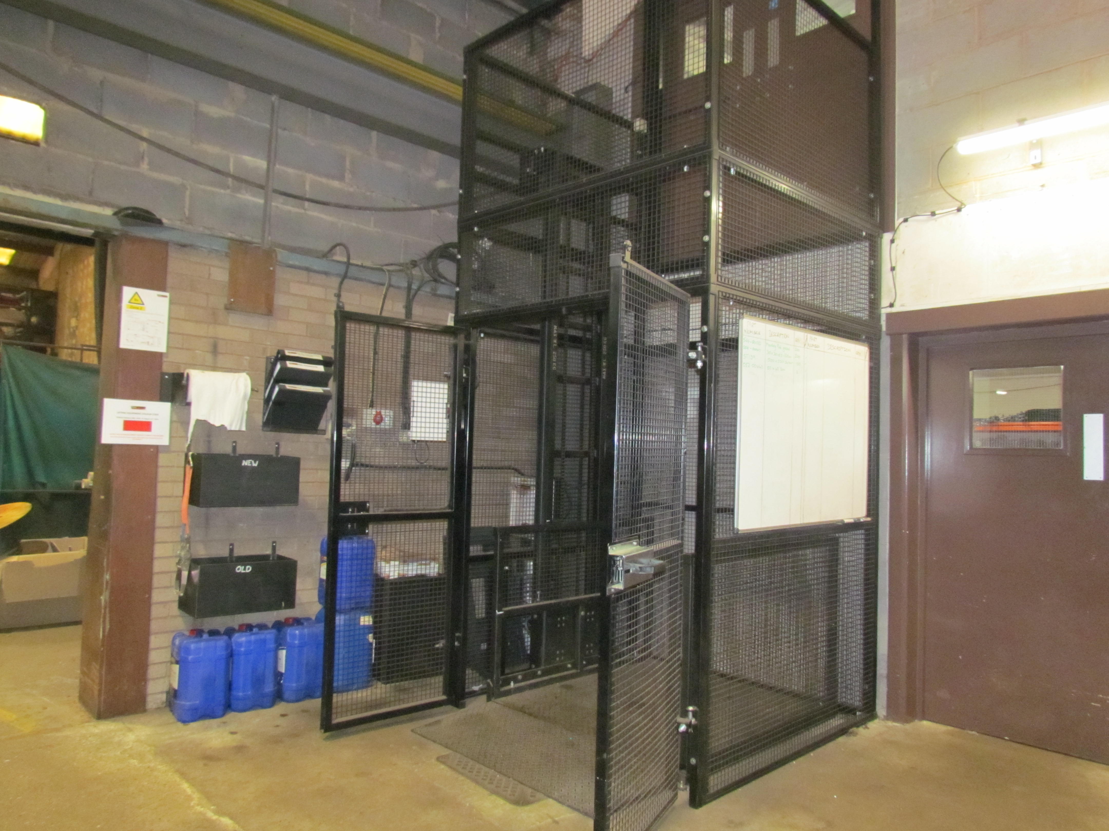 A black metal cage lift with mesh panels is situated in an industrial warehouse. Blue containers and hanging clipboards are nearby, and a whiteboard is mounted on the wall next to a brown door.