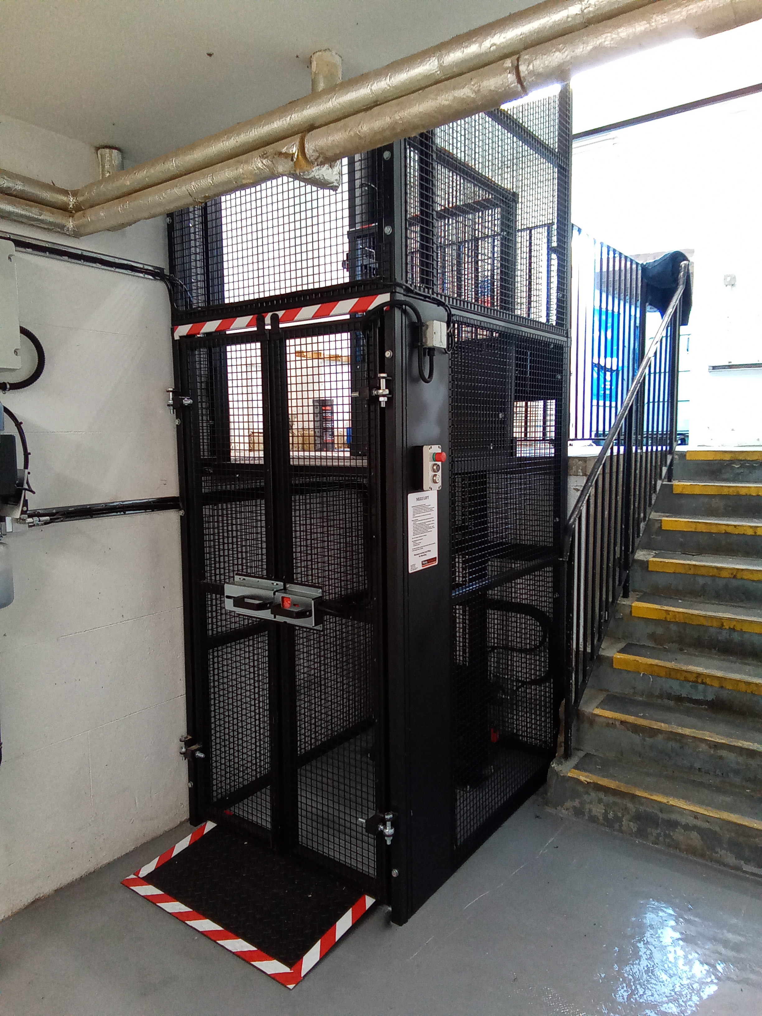 A black metal platform lift with mesh sides is installed beside a staircase in an industrial indoor setting. Yellow-striped steps are next to the lift, which has warning stripes on the platform edges.