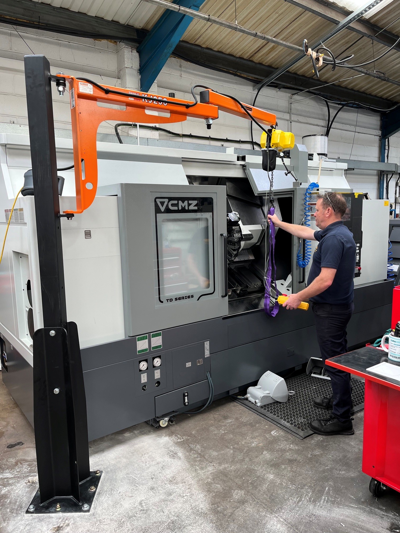 A man operates a yellow industrial hoist to lift or load parts into a large CMZ-branded CNC machine in a workshop setting.