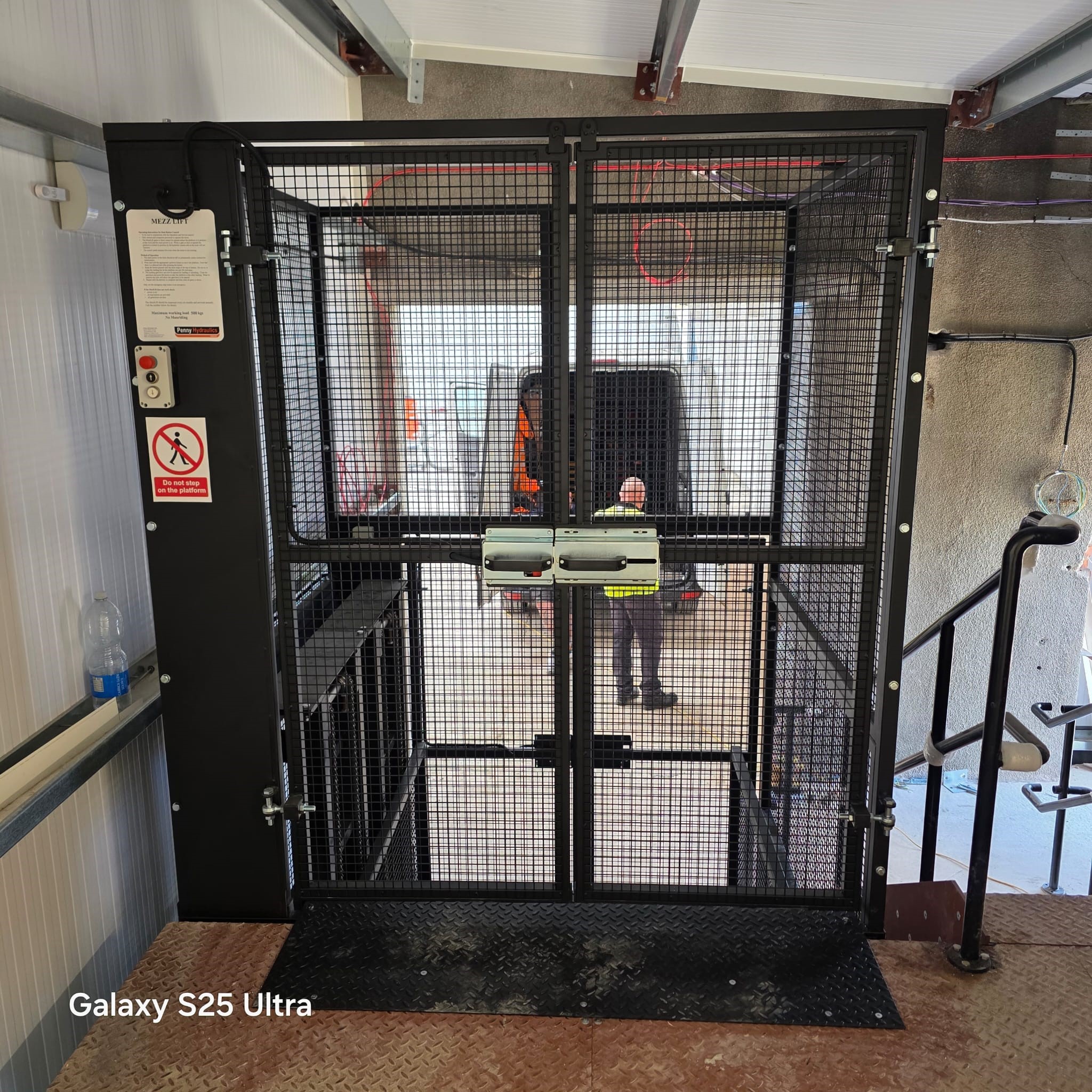 A black metal lift cage with mesh sides is positioned at the edge of an industrial platform. A person in safety gear is visible in the background through the cage. A red emergency button and safety instructions are on the left panel.