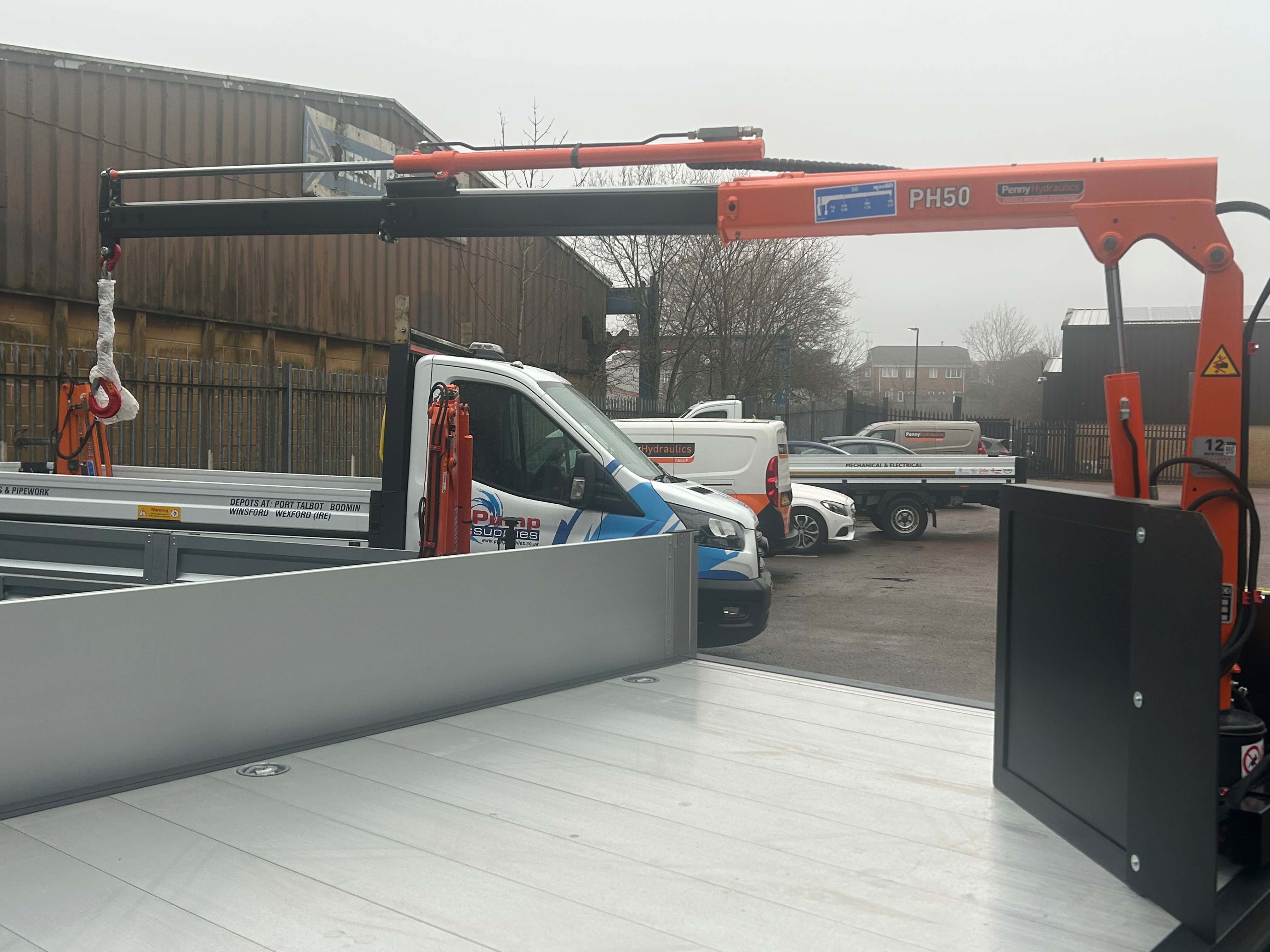 A small orange crane labelled PH50 is mounted on a flatbed lorry, with its arm extended. Several white vehicles and industrial buildings are visible in the background.