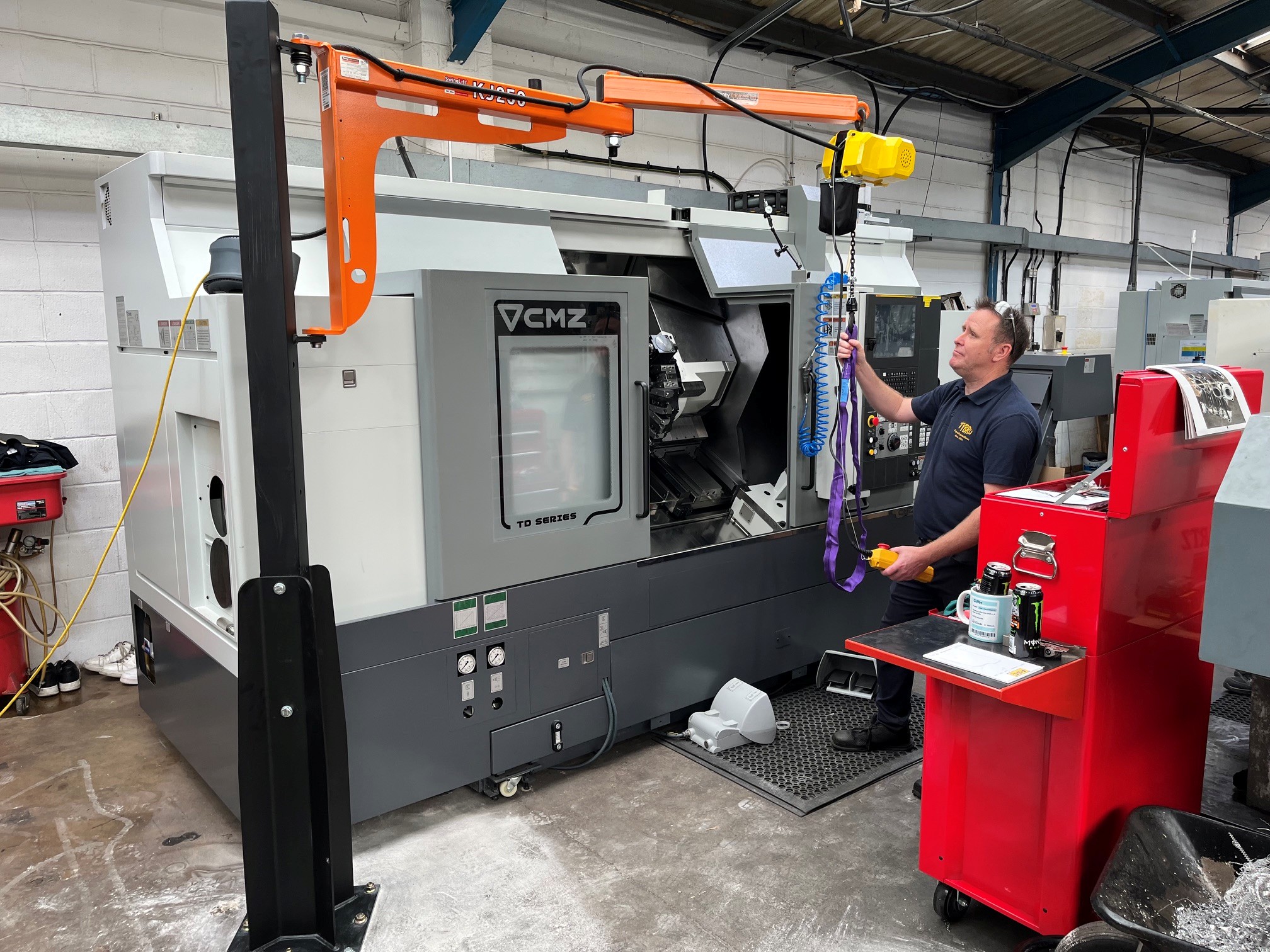 A man operates a yellow overhead hoist near a large industrial CNC machine in a workshop, with tools and a red trolley nearby.