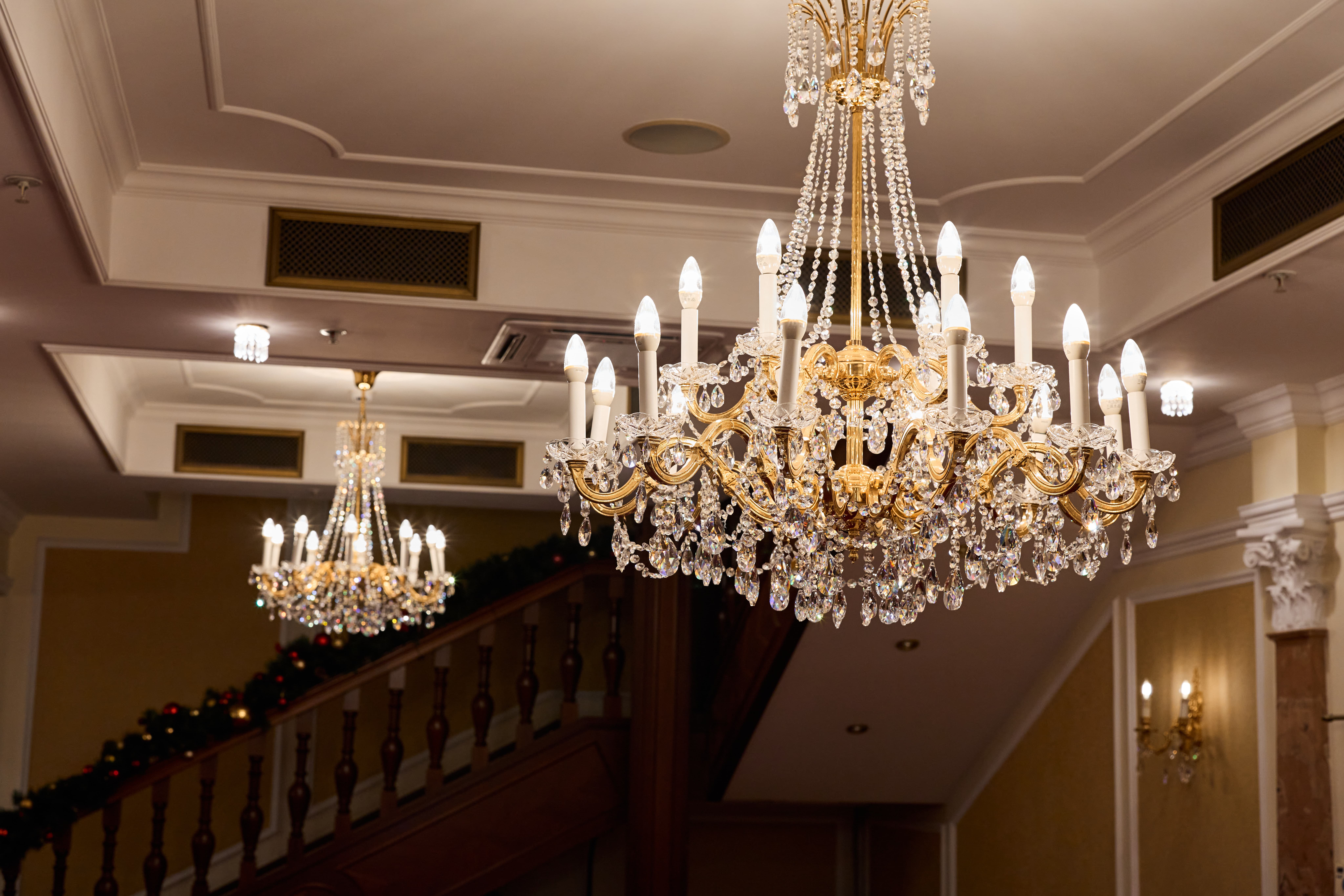 A close-up view of ornate, sparkling crystal chandeliers hanging from a high, elegant ceiling in a grand interior space. The image is partially distorted with digital glitches.