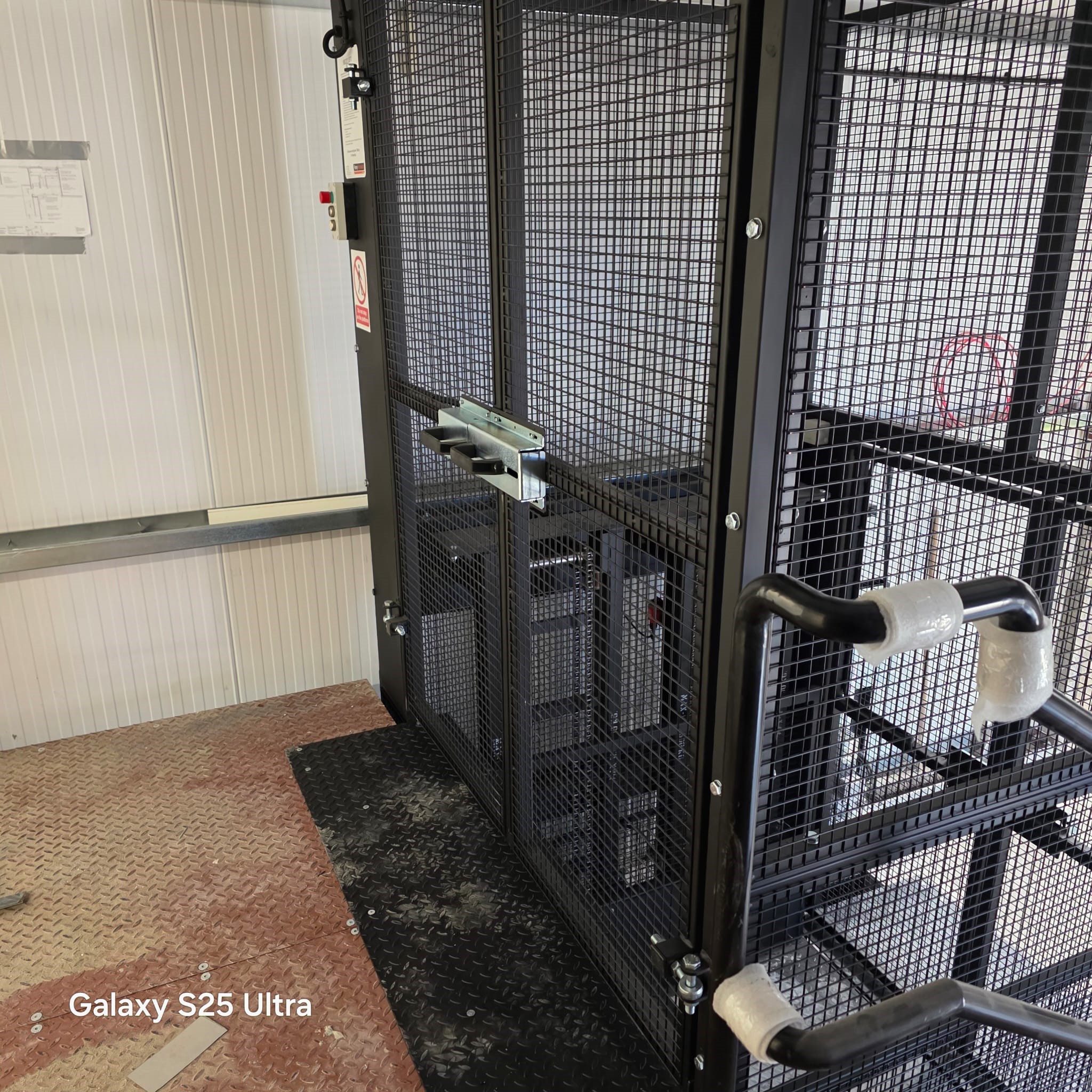 A black metal safety cage with mesh panels and a secure latch is situated on a textured floor beside a white corrugated wall. Handrails with grey padding are visible in the foreground. Galaxy S25 Ultra text appears in the corner.