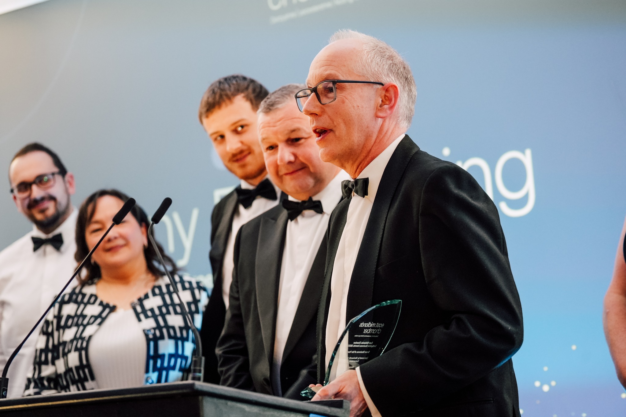 A group of people in formal attire stand together on stage. A man in front, holding an award, speaks at a lectern while others smile behind him. A blue backdrop with white text is visible.