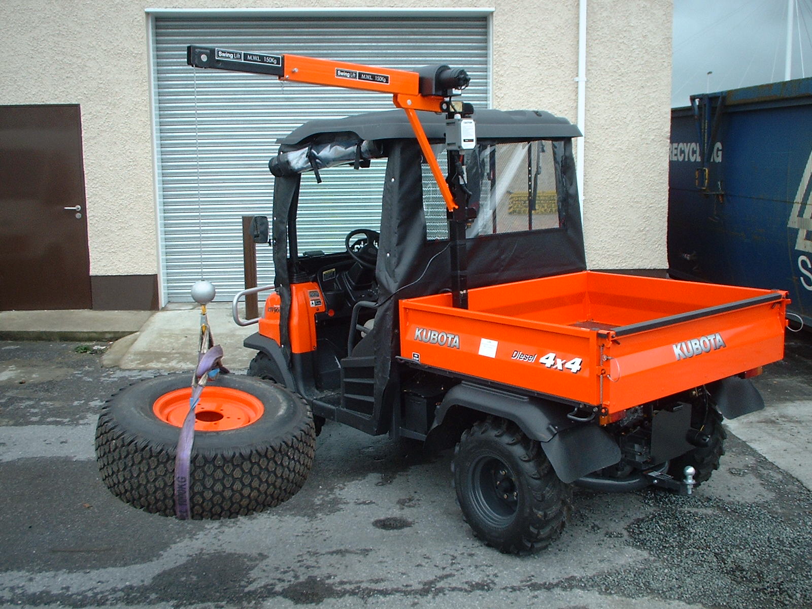 An orange Kubota 4x4 utility vehicle with a small crane attached to the back is lifting a large tyre with a purple strap in an industrial area near a building with a metal shutter door.