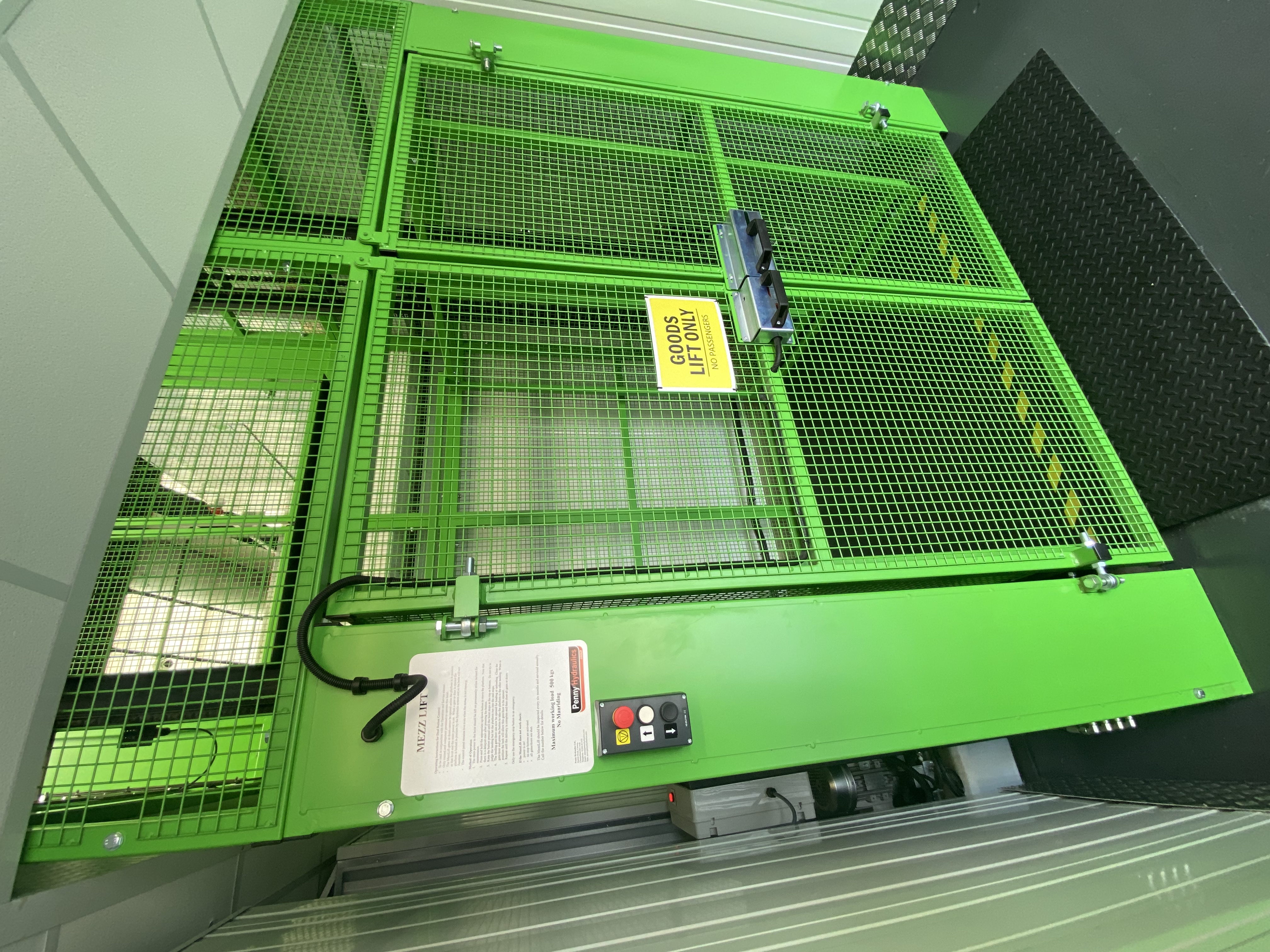 A green industrial goods lift with mesh doors and safety signs stands in a warehouse. A control panel is mounted on the left side, and a yellow sign on the door reads GOODS LIFT ONLY.