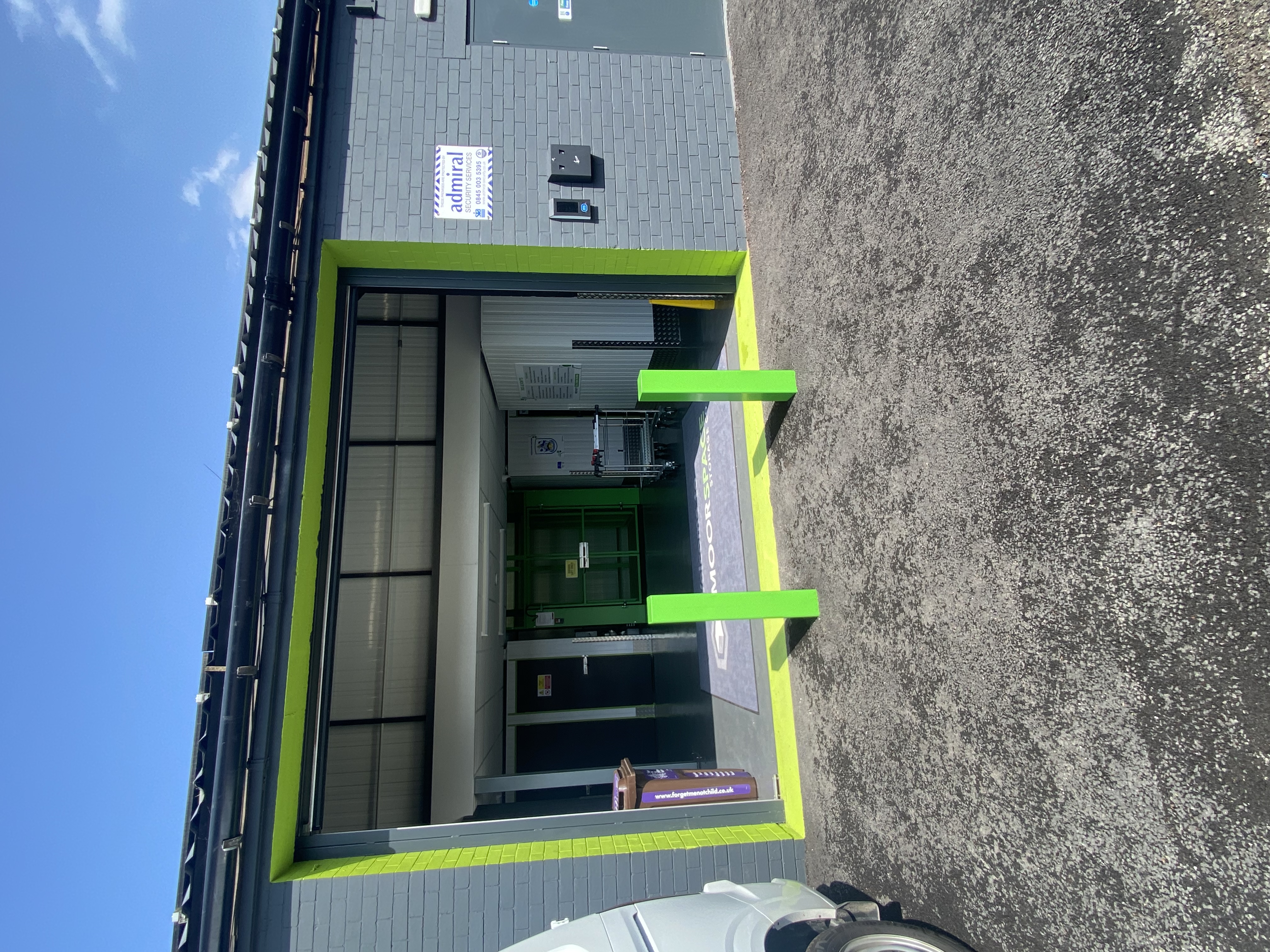 A storage facility entrance with bright green trim, bollards, and an open roller shutter. A van is parked on the left, and a sign reads admiral. Shopping trolleys are visible inside the building.