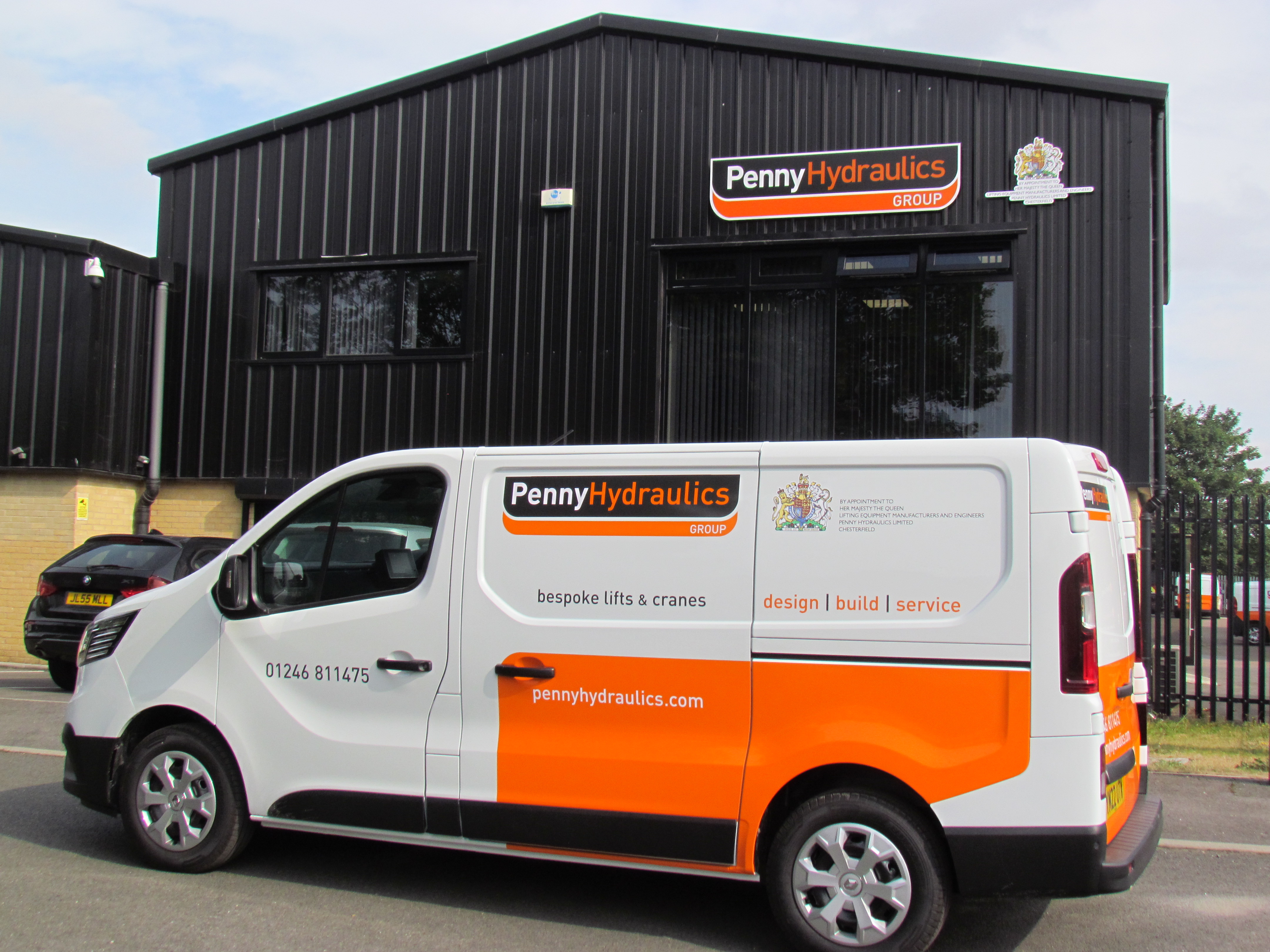 A white and orange Penny Hydraulics van is parked outside a black industrial building with the Penny Hydraulics Group logo. The van advertises bespoke lifts and cranes, with contact details and website on its side.