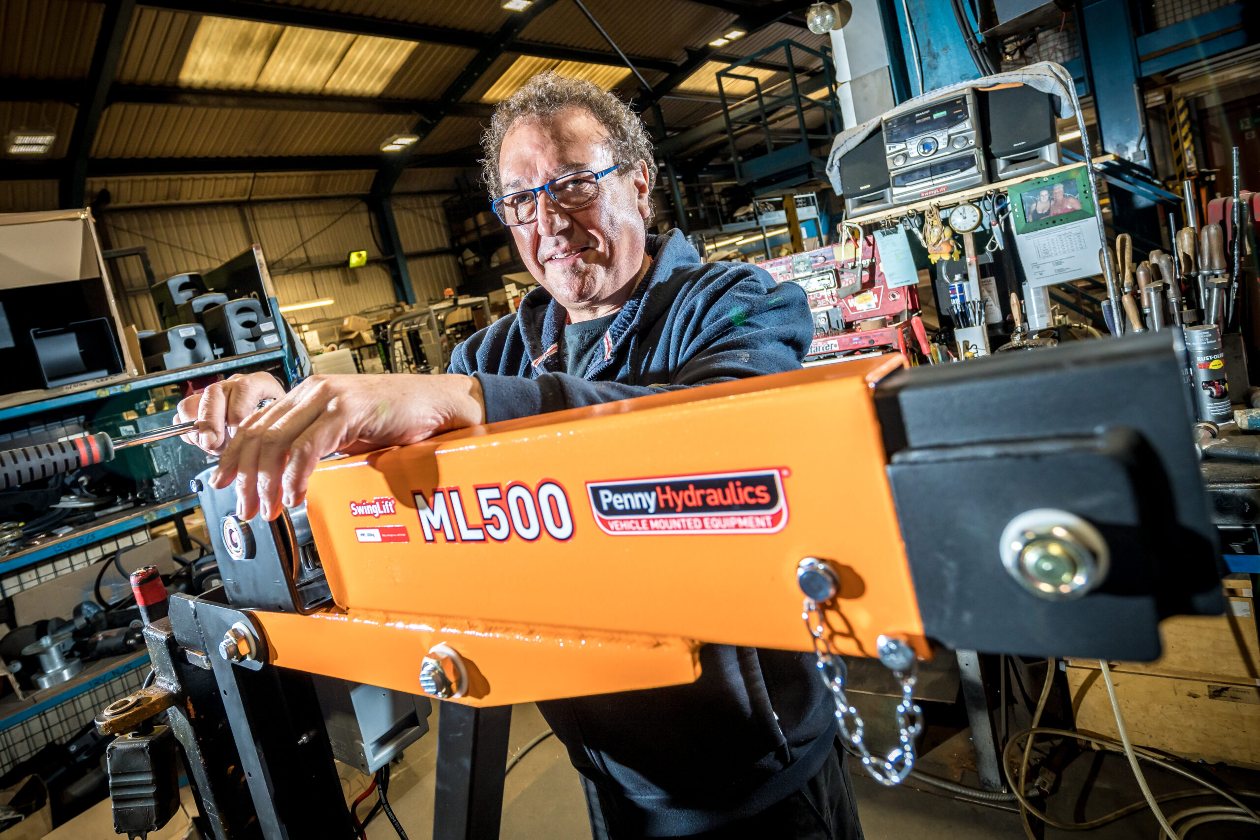 A person wearing glasses stands in a workshop, holding a large orange hydraulic lifting arm labelled ML500 Penny Hydraulics. Tools, machinery, and shelves are visible in the background.