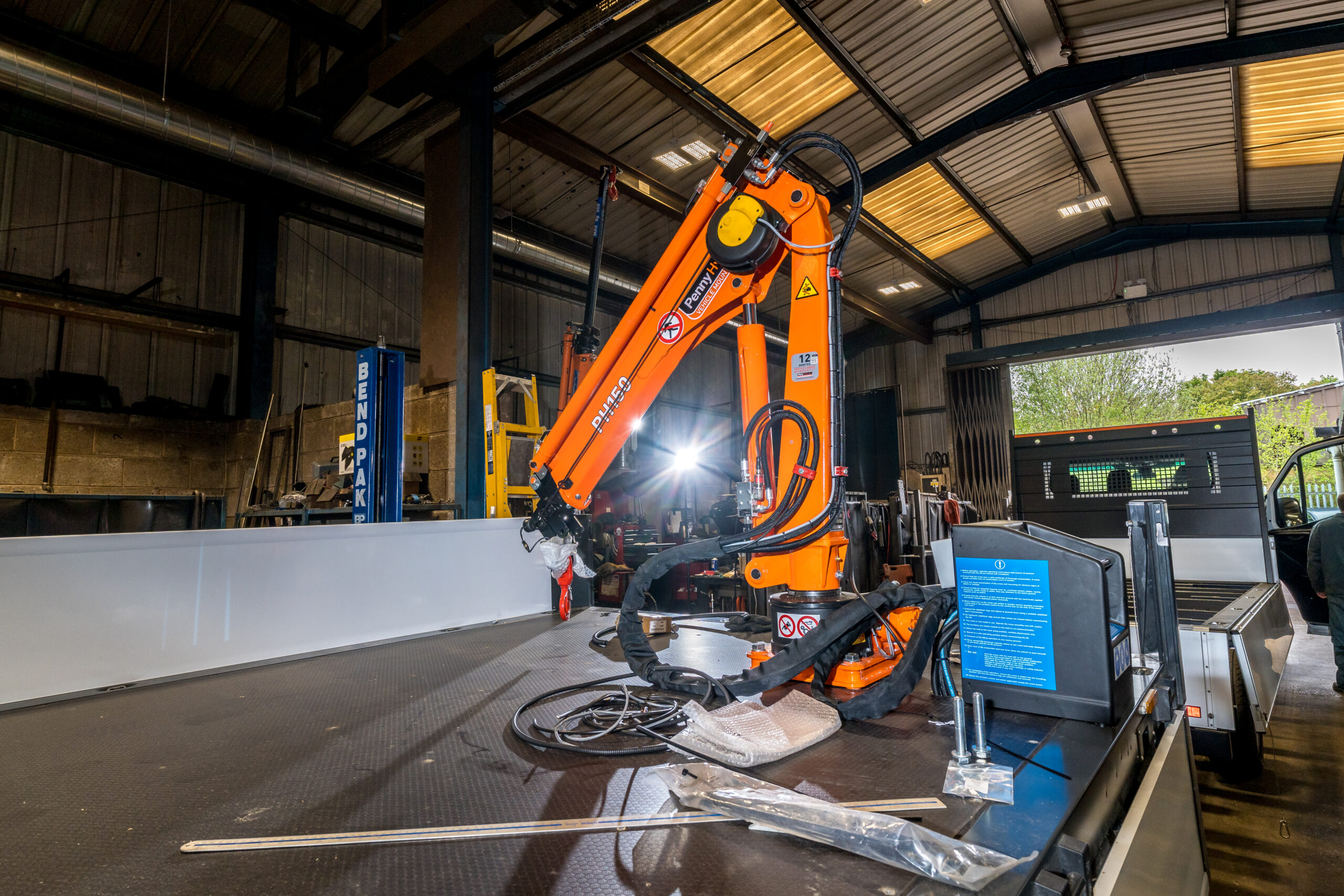 An orange industrial robotic arm is mounted on a metal platform inside a warehouse. Tools, cables, and equipment are scattered on the work surface. Sunlight filters through the windows in the background.