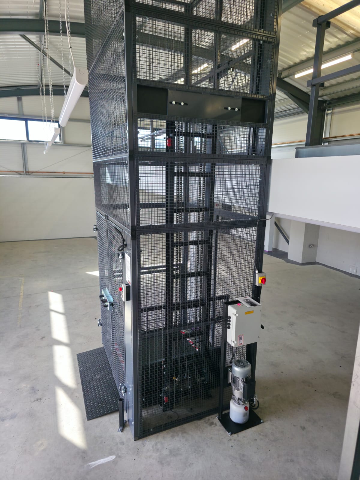 A modern glass lift shaft inside a building, with visible black metal framework and glass panels. The surrounding area features structural beams and light streaming in from the windows.