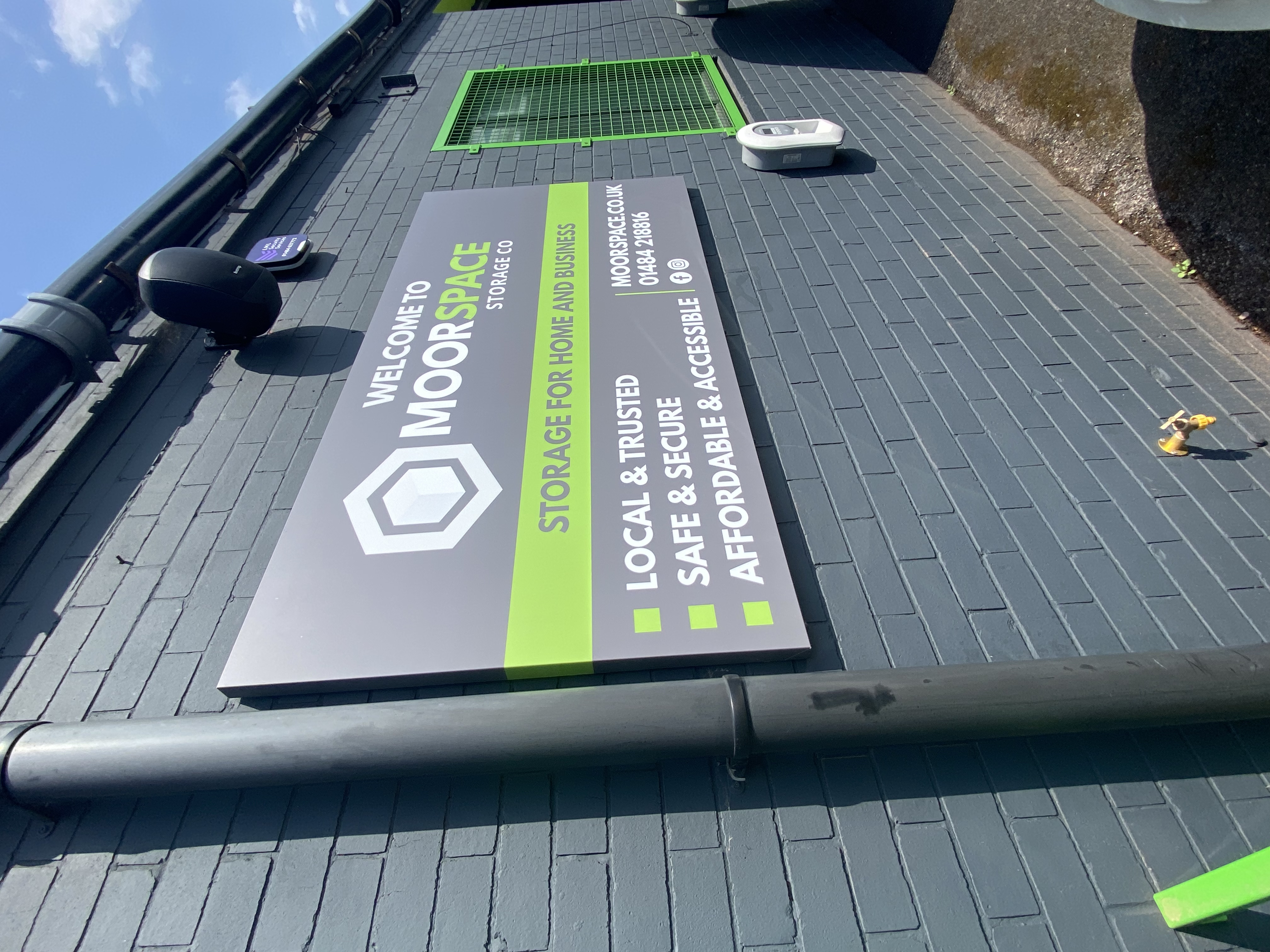 A grey brick wall displays a MoorSpace Storage sign promoting secure, affordable, and accessible storage for home and business. A small green-trimmed window and black drainpipe are also visible.