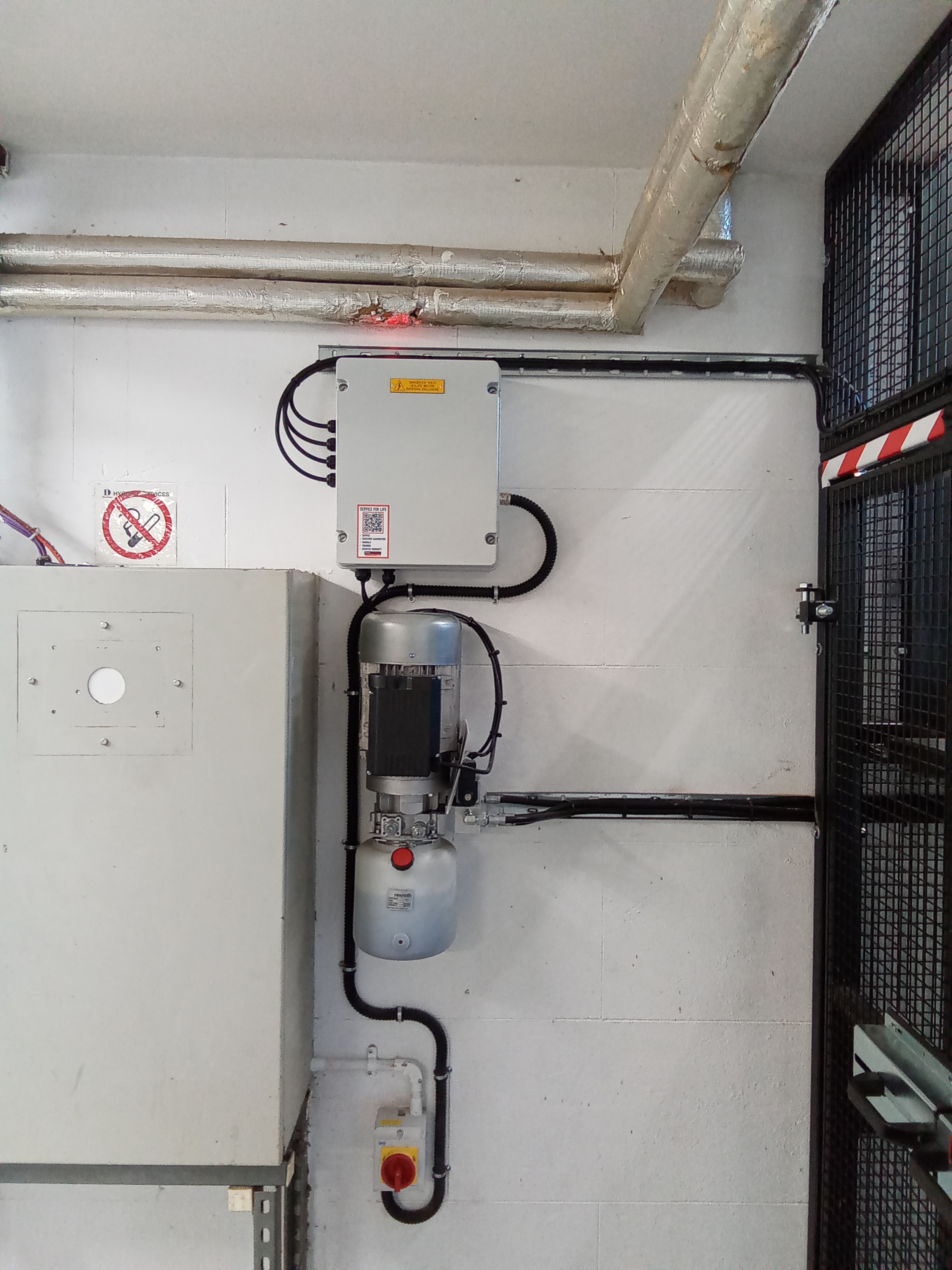 An electrical control box and a pump system mounted on a white wall, with connected pipes and cables. A “no smoking” sign is visible on the wall above a large grey metal panel.