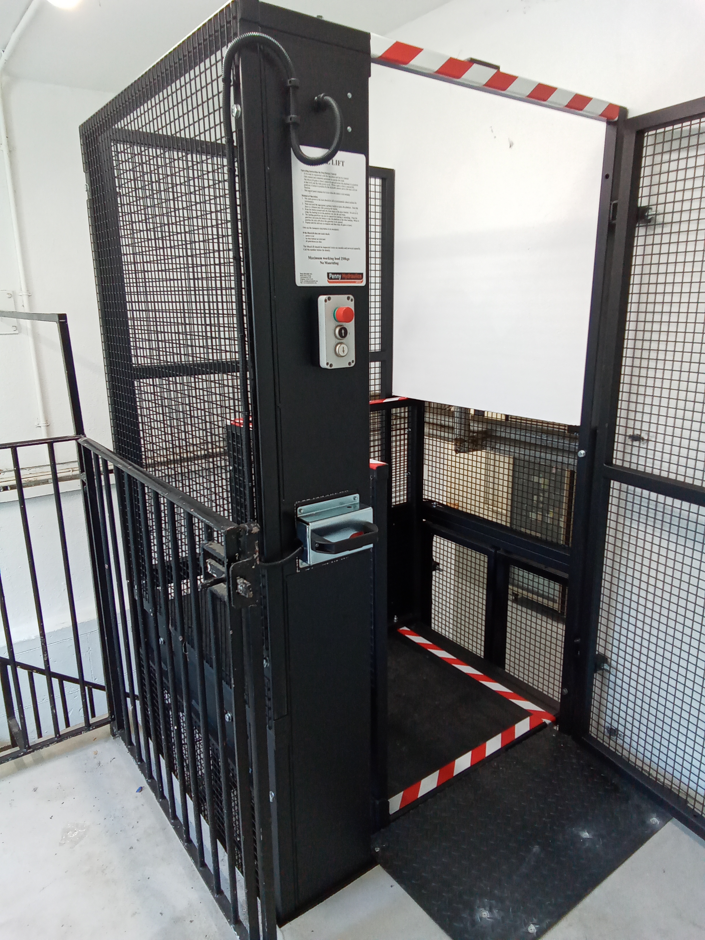 A black metal platform lift with safety railings and a mesh enclosure, used for accessibility. The lift has red and white caution stripes, an emergency stop button, and an instruction sign posted on the control panel.