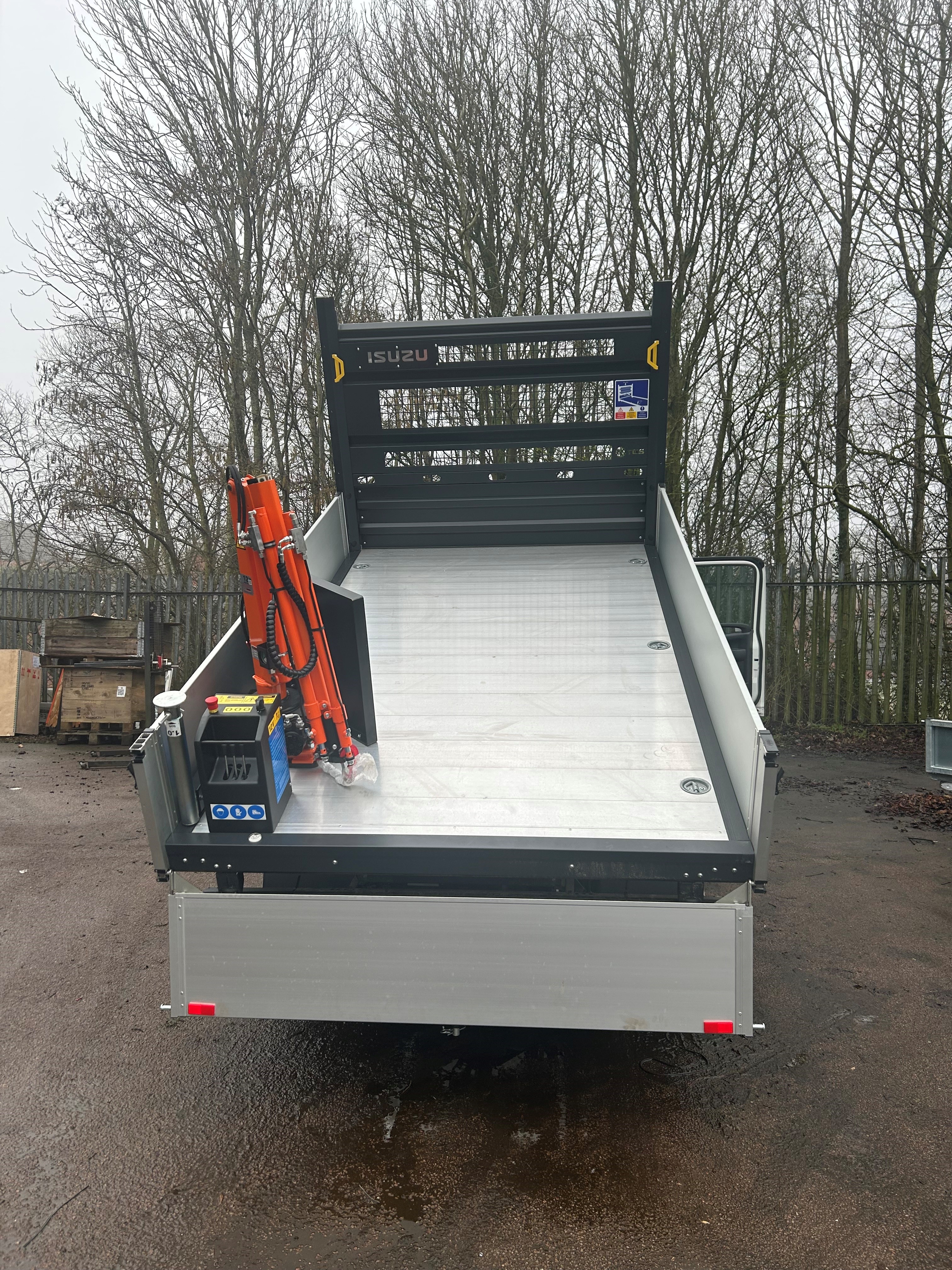 A rear view of a tilted flatbed Isuzu lorry with an orange crane arm on the left side, parked outdoors on wet pavement, with trees in the background.