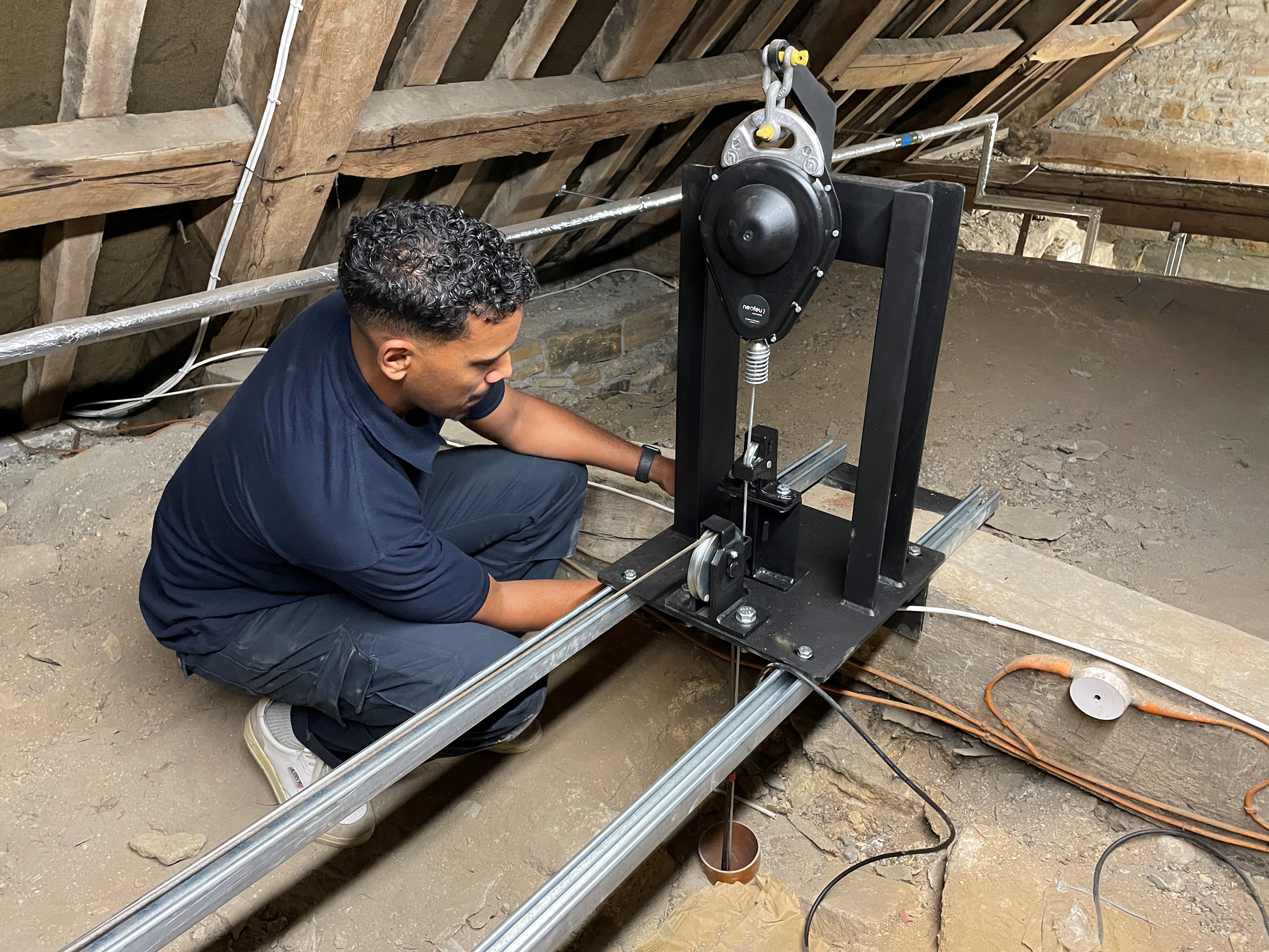 A man kneels in a loft, adjusting a mechanical device mounted on metal rails. The area is dusty, with exposed beams, wires, and electrical leads visible around him.