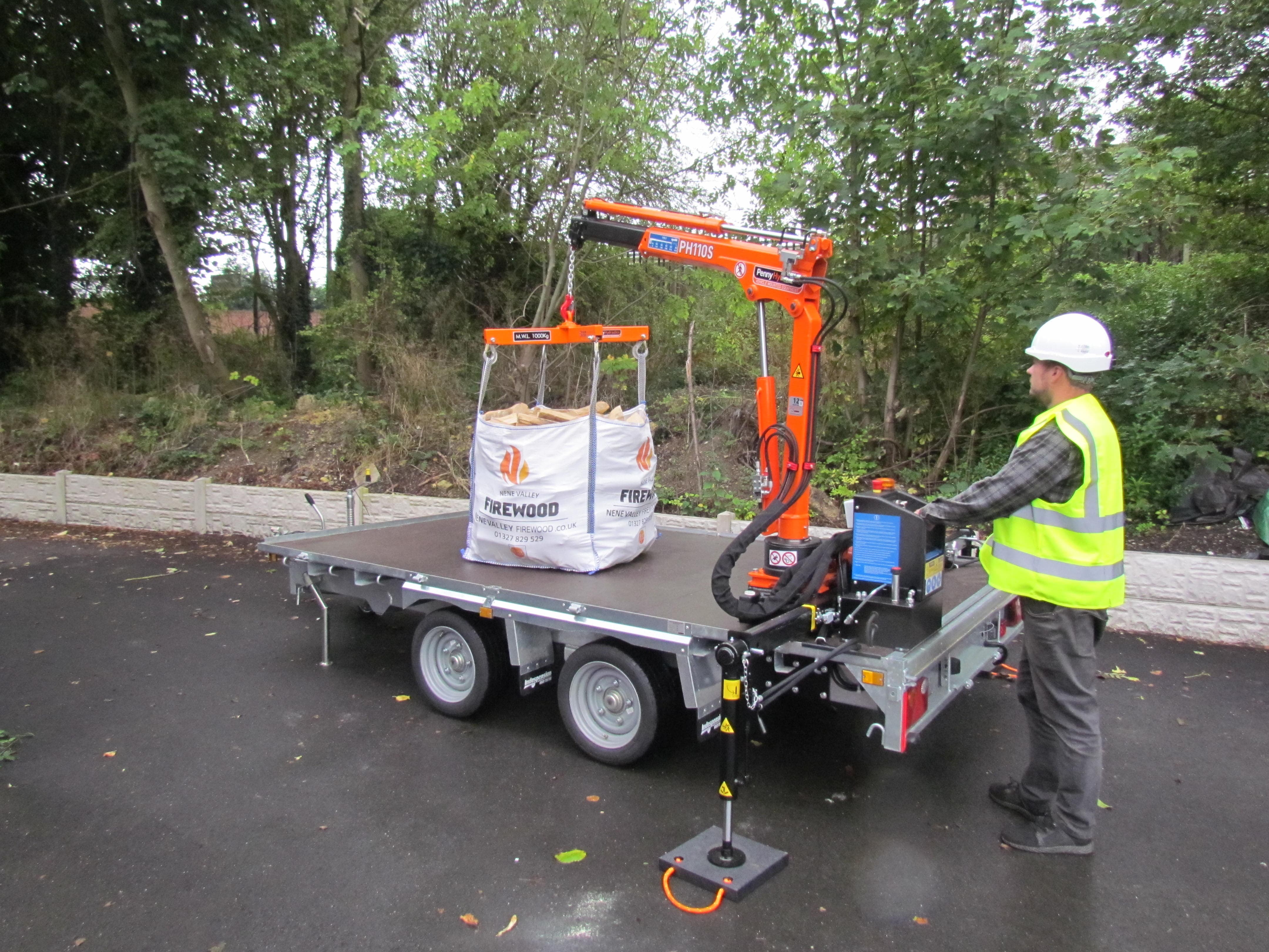 A person in a yellow reflective vest stands next to a small trailer loaded with white bags or boxes on a paved path surrounded by green trees. The image is distorted with digital glitches.