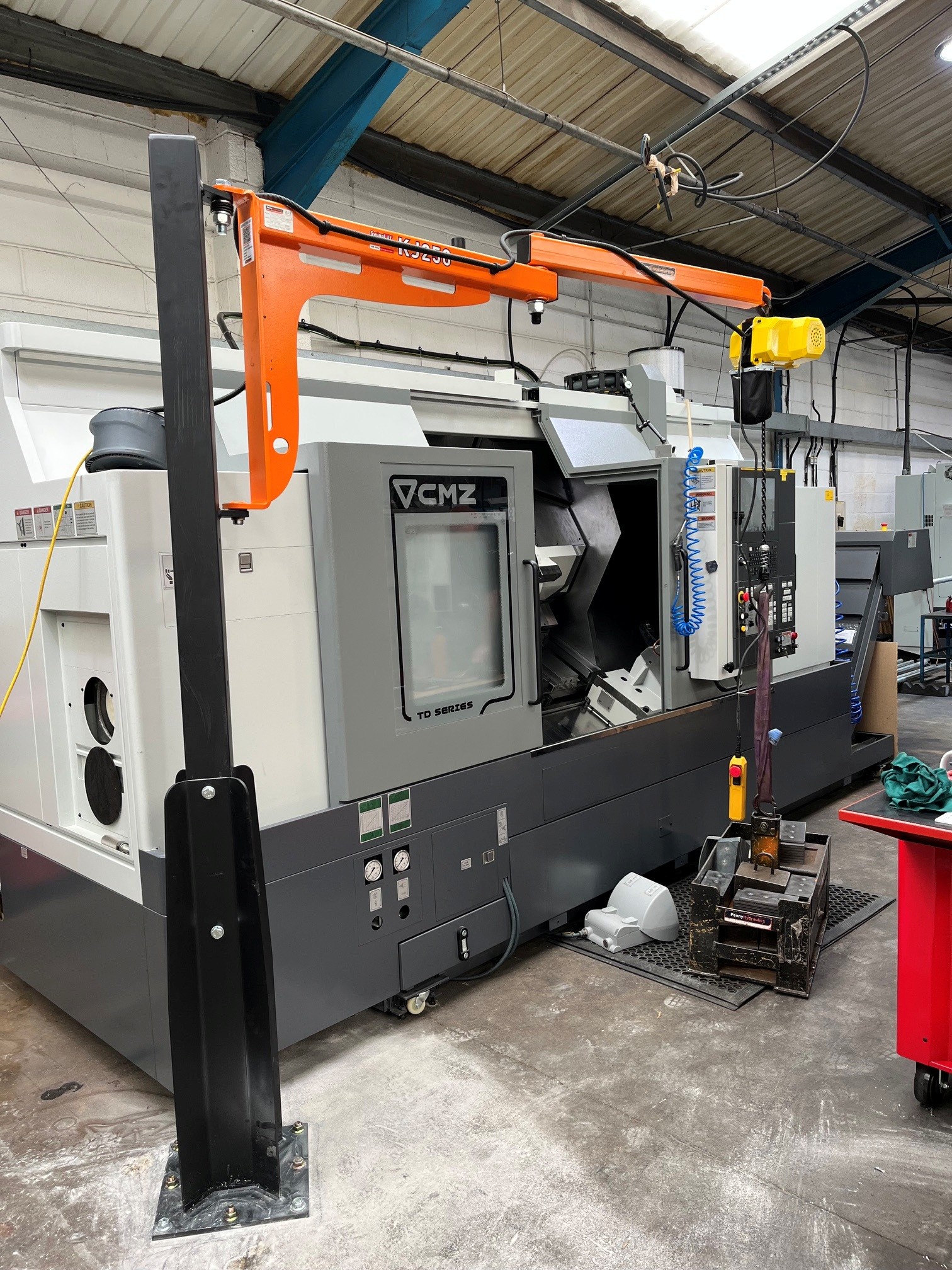 A CNC machine sits in an industrial workshop with an orange gantry crane and yellow hoist attached, used for lifting heavy parts. The area is clean and organised.
