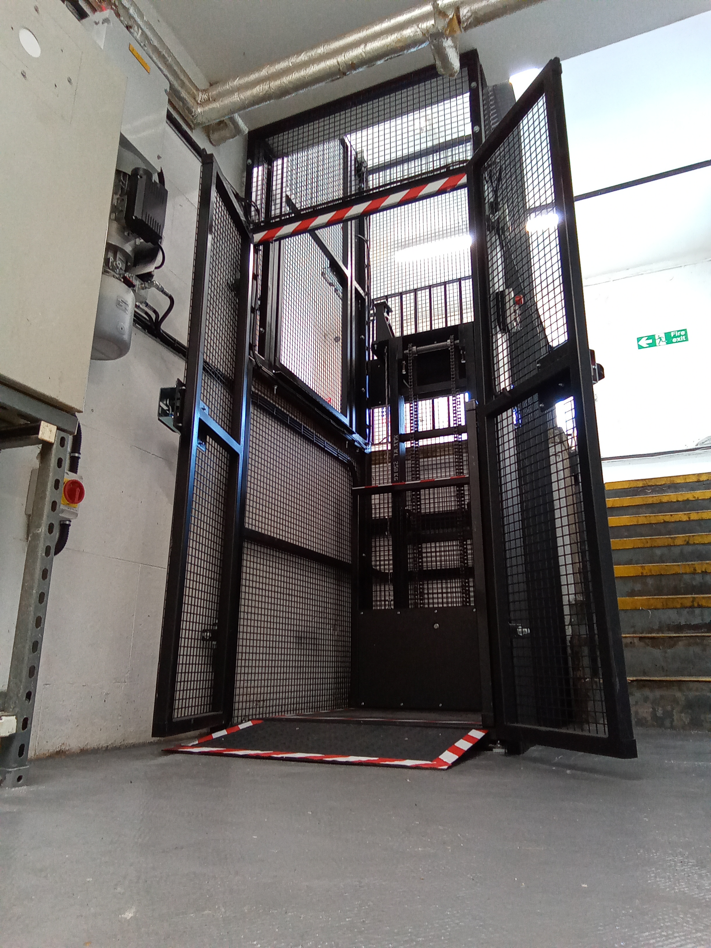 A metal platform lift with safety gates and mesh sides stands open in an industrial corridor, next to a flight of yellow-striped stairs and control panels mounted on the white wall.