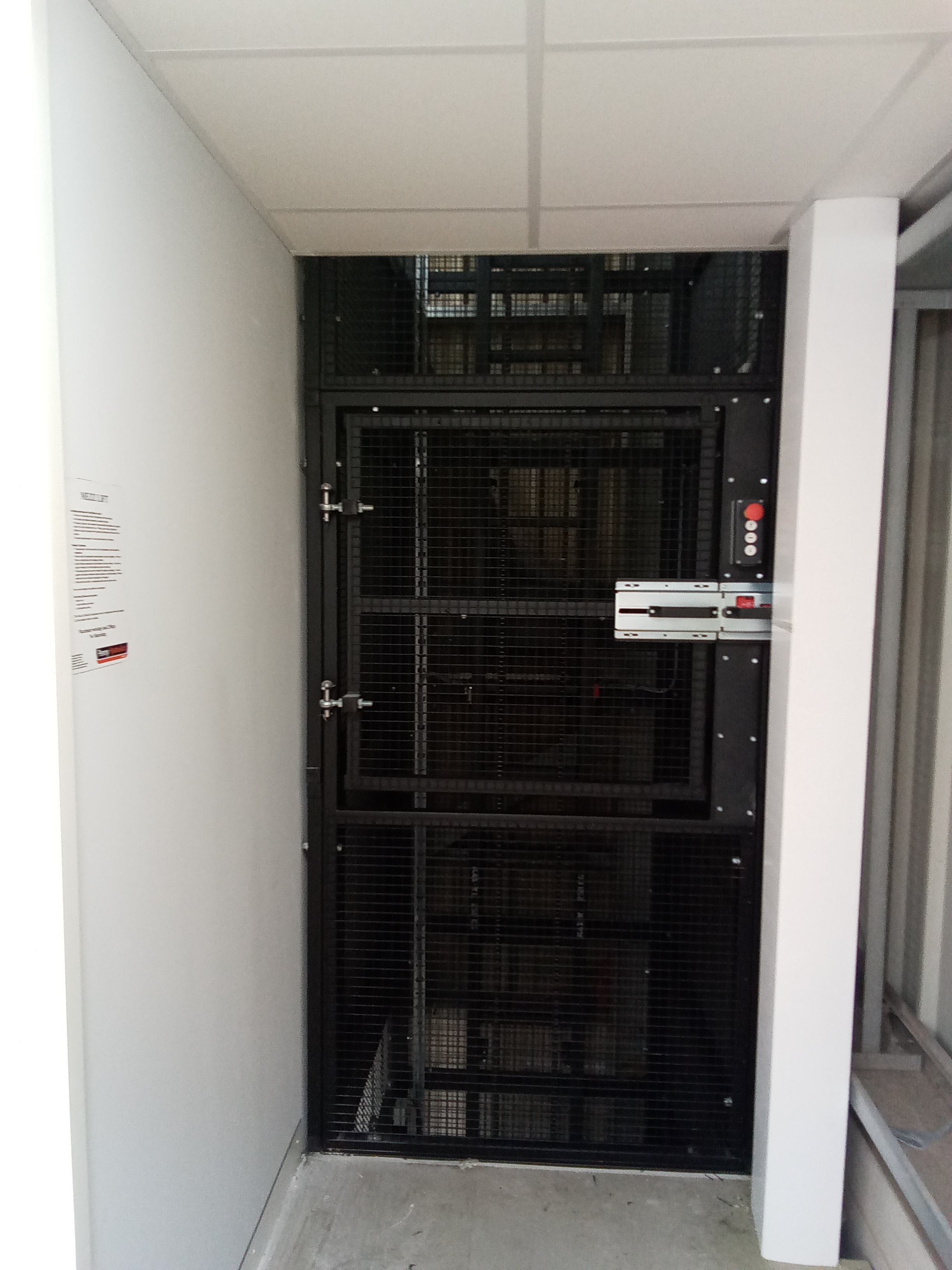 A black metal security gate with a keypad and electronic lock blocks access to a stairwell in a corridor with white walls and a tiled ceiling. A notice is posted on the wall to the left of the gate.