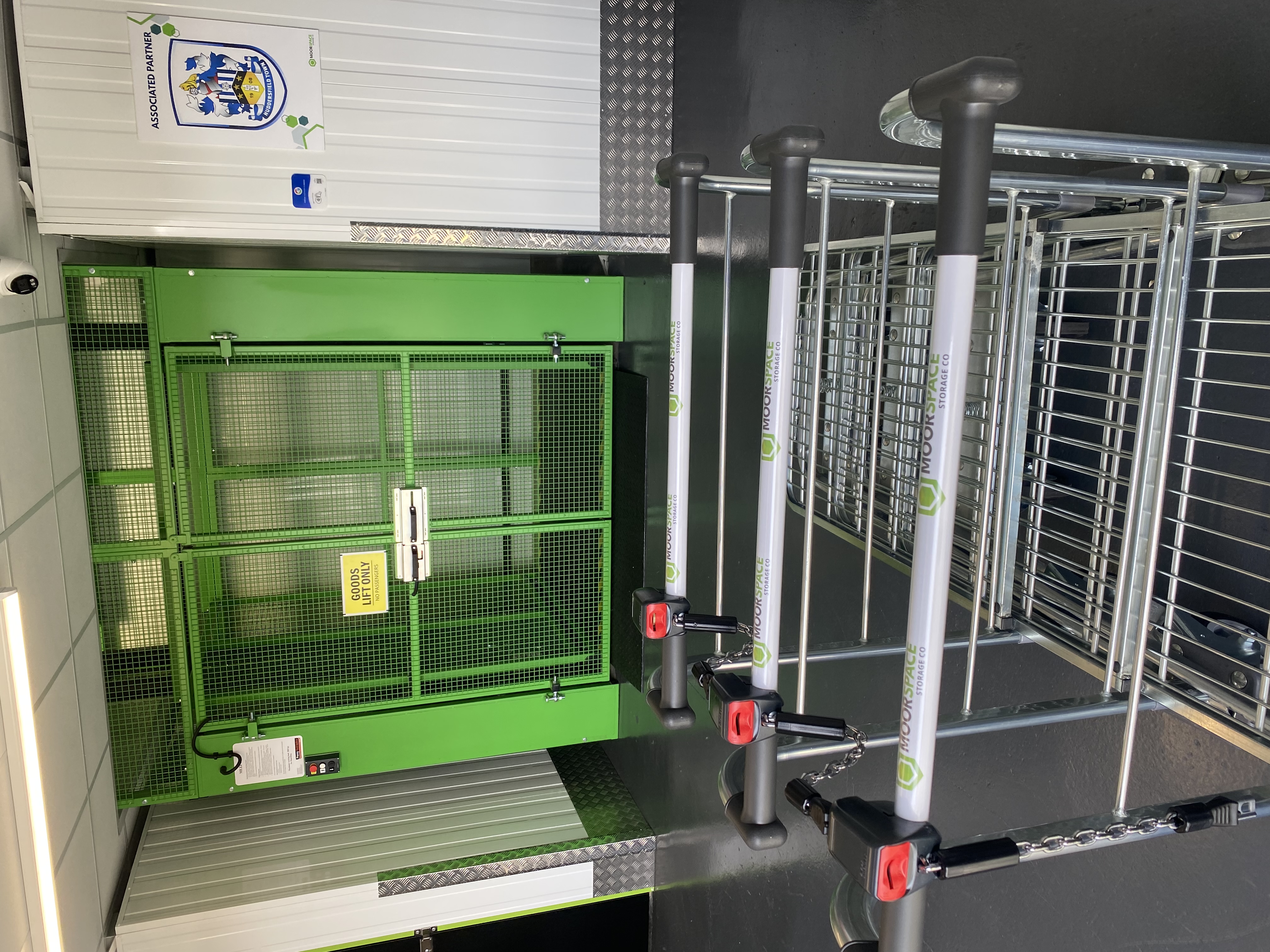 Three empty trolleys are in front of a green secure goods collection area with a yellow Goods Lift Only sign, inside a modern, well-lit facility.