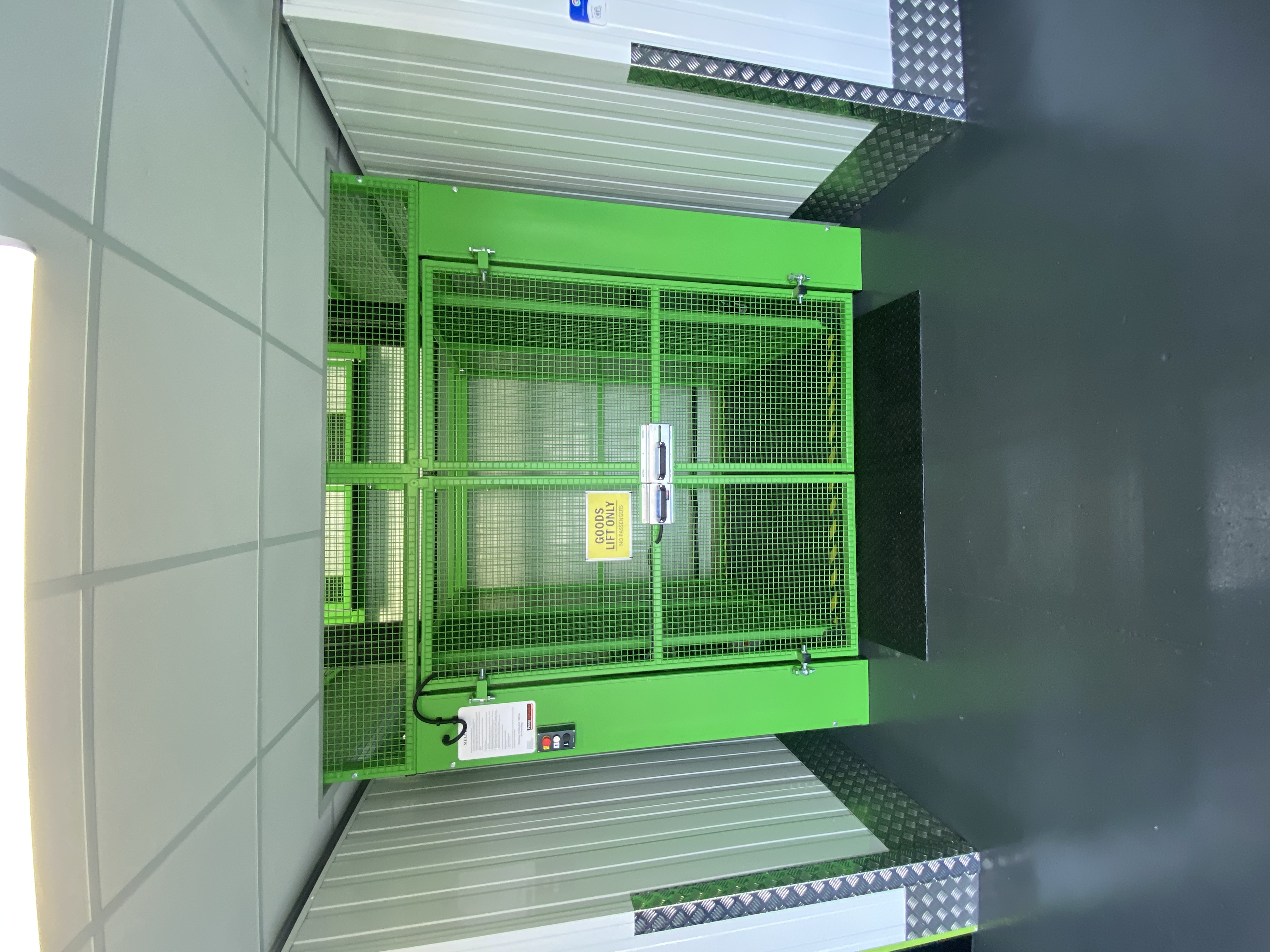 A green metal cage lift with a yellow Goods Only sign stands in a corridor with grey floors and white walls. The ceiling is tiled, and a black mat is in front of the lift doors.