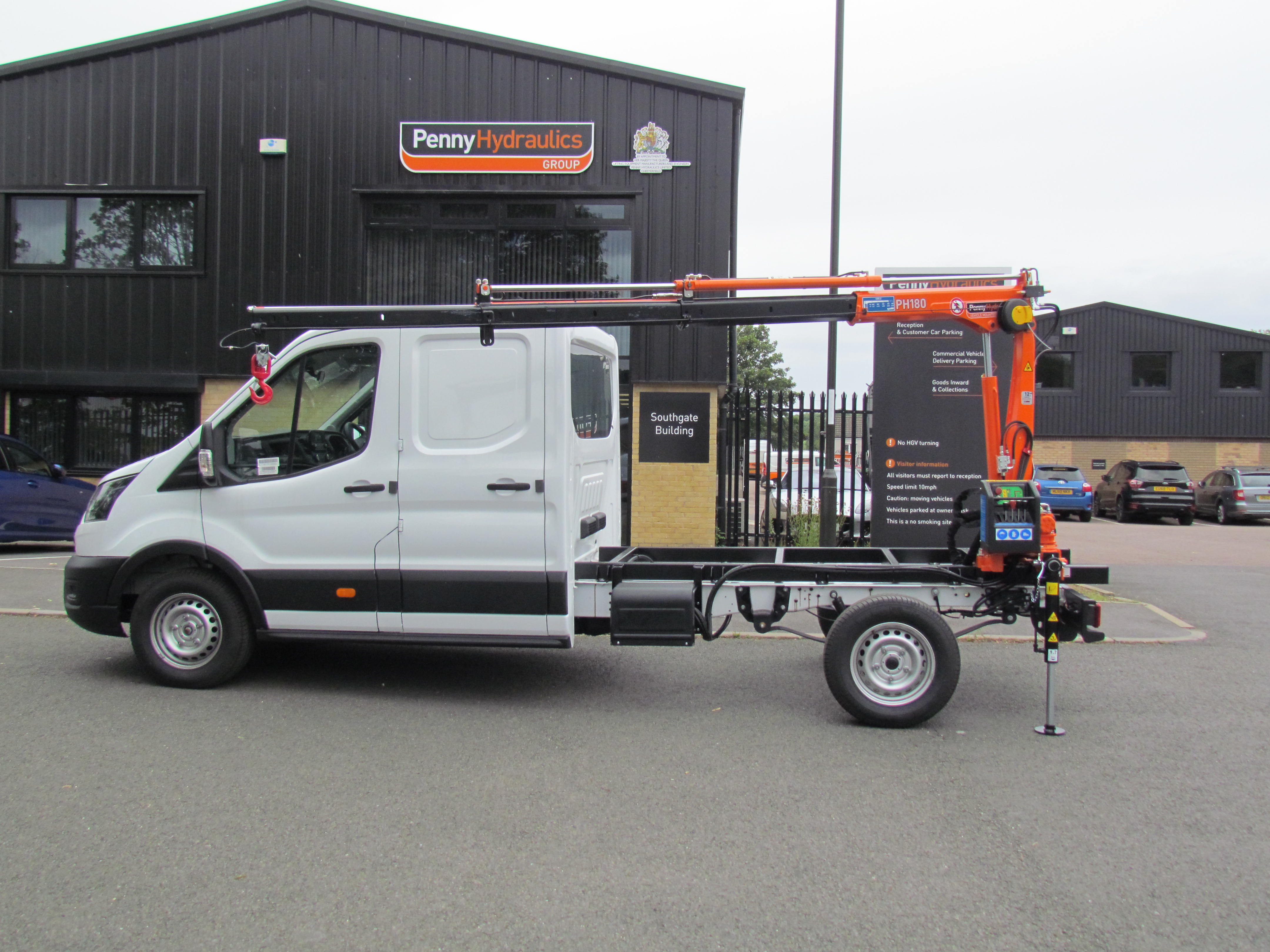 A white utility van with an orange hydraulic crane mounted on its rear is parked outside a black industrial building with a sign reading Penny Hydraulics. Other vehicles and buildings are visible in the background.