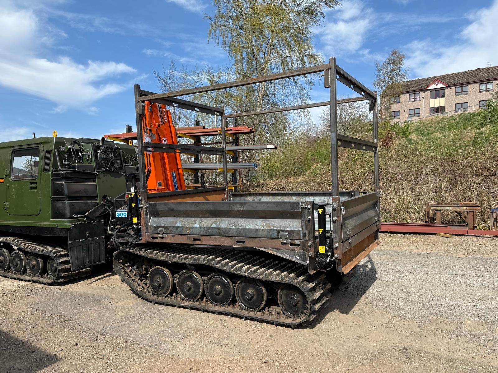 A green tracked vehicle with an open metal cargo bed and frame is parked outdoors near some orange equipment, with trees and a building in the background under a partly cloudy sky.