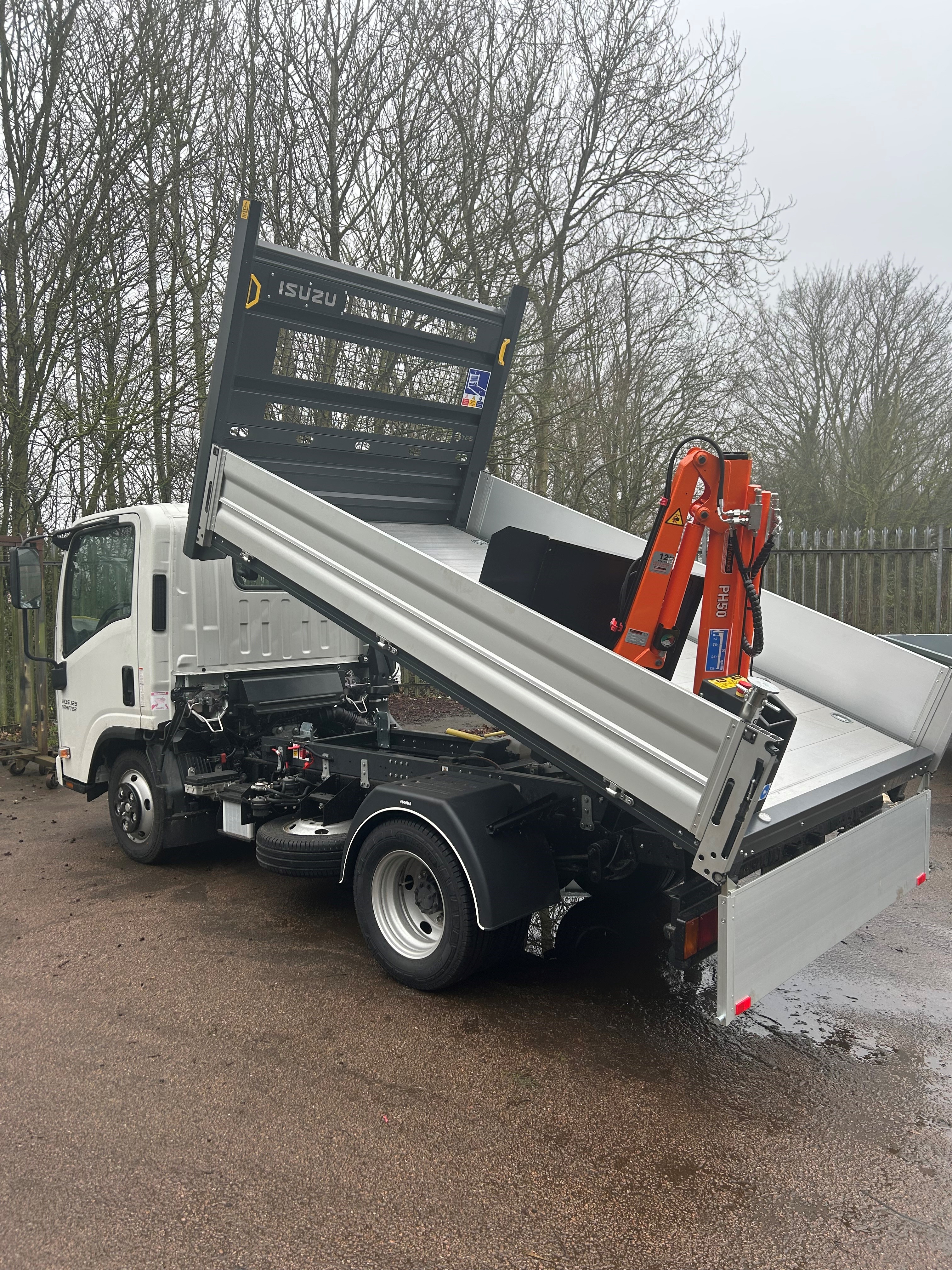 A white flatbed lorry with its bed tilted upward, equipped with a small orange crane, is parked on wet tarmac near leafless trees and a metal fence on a cloudy day.