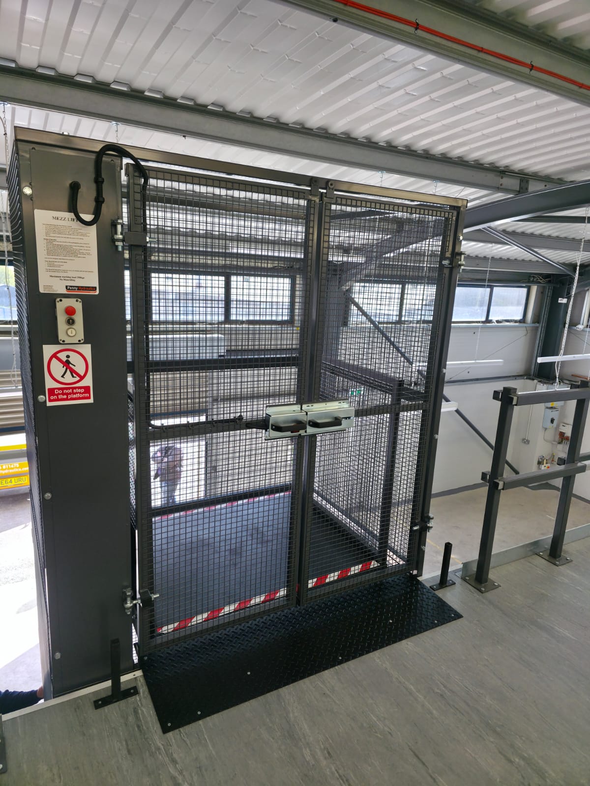 A city bus stopped at a bus shelter. The shelter has a bench and glass walls, and there is a person waiting inside. The bus doors are open, and a street is visible in the background.
