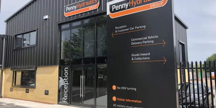 A distorted image showing a modern building with a slanted roof, a large entrance, and a black vertical sign in front. The image has pixelated lines and artefacts, making the text and details difficult to read.