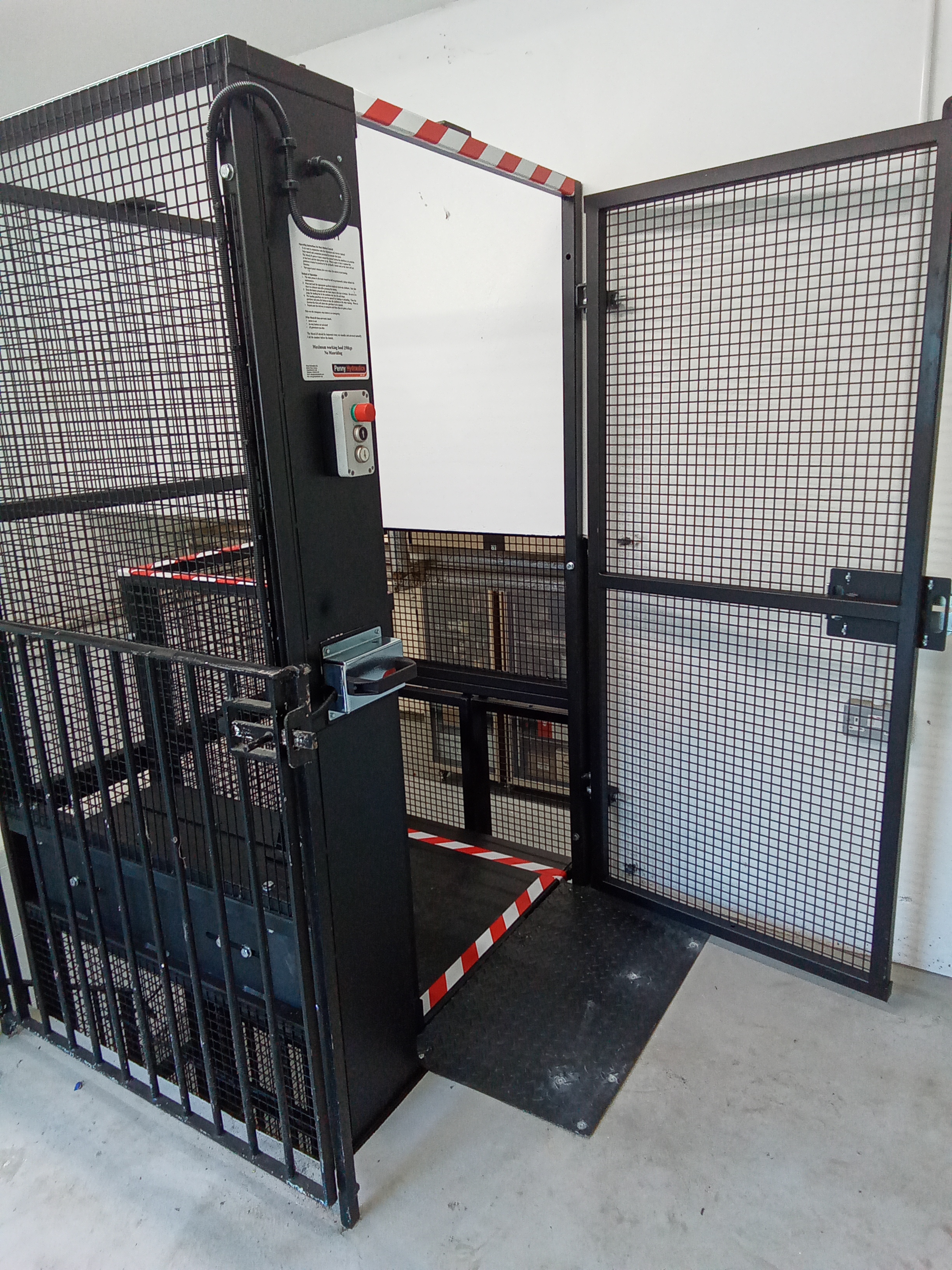 A black metal cage lift with an open mesh door and safety stripes, located indoors on a concrete floor. The lift has control buttons and an instruction panel on the side.